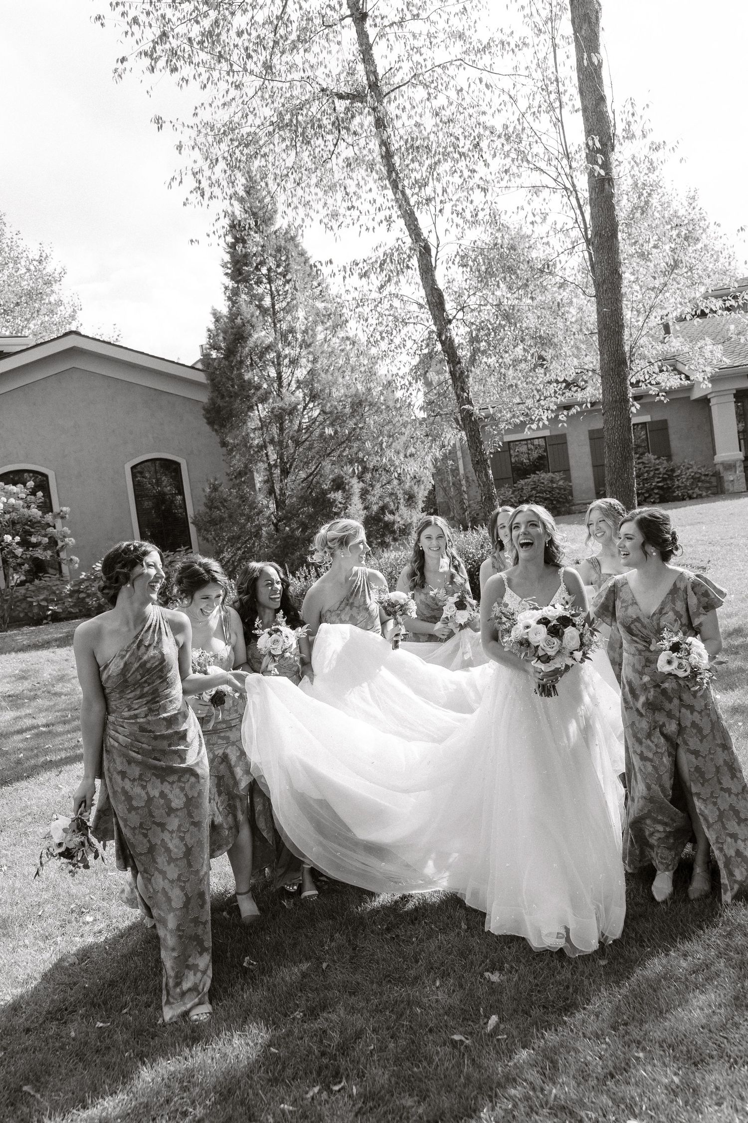 Black and white photo of bridesmaids in floral dresses helping with a bride's flowing dress outdoors.