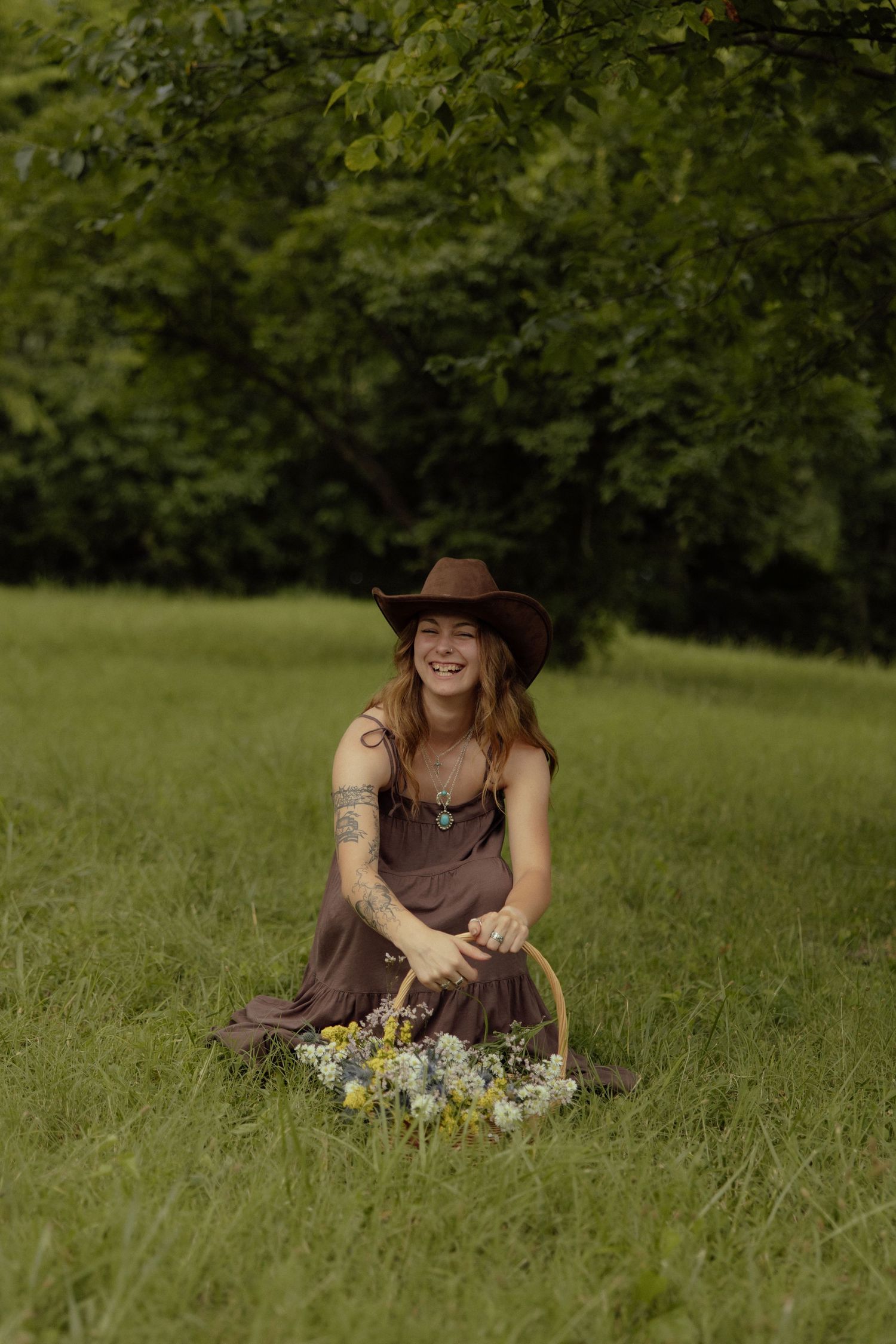 A person in a brown dress and hat sits in a grassy meadow with wildflowers on a sunny summer day.