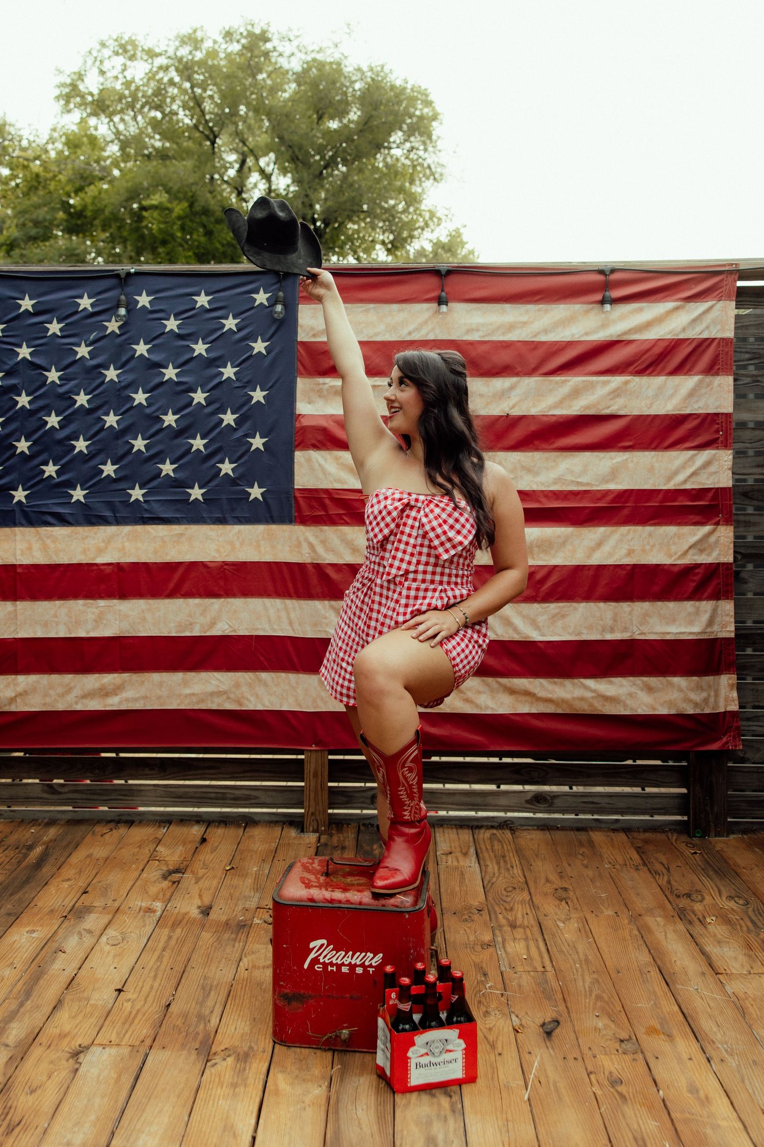 A patriotic scene with red and white gingham dress and cowboy boots against an American flag backdrop on wooden deck.