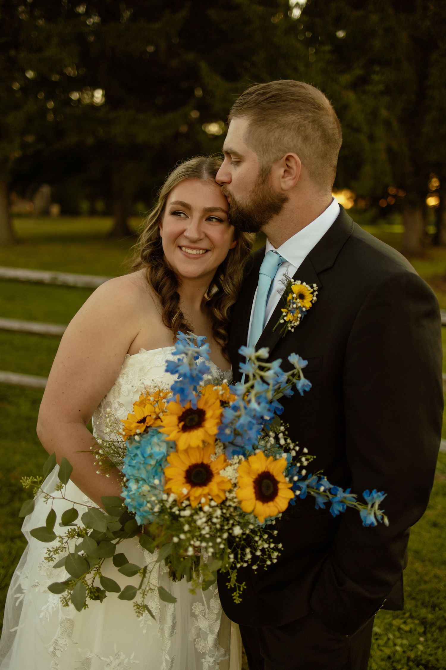 A romantic wedding couple embraces in a garden setting with a vibrant sunflower and blue delphinium bouquet.