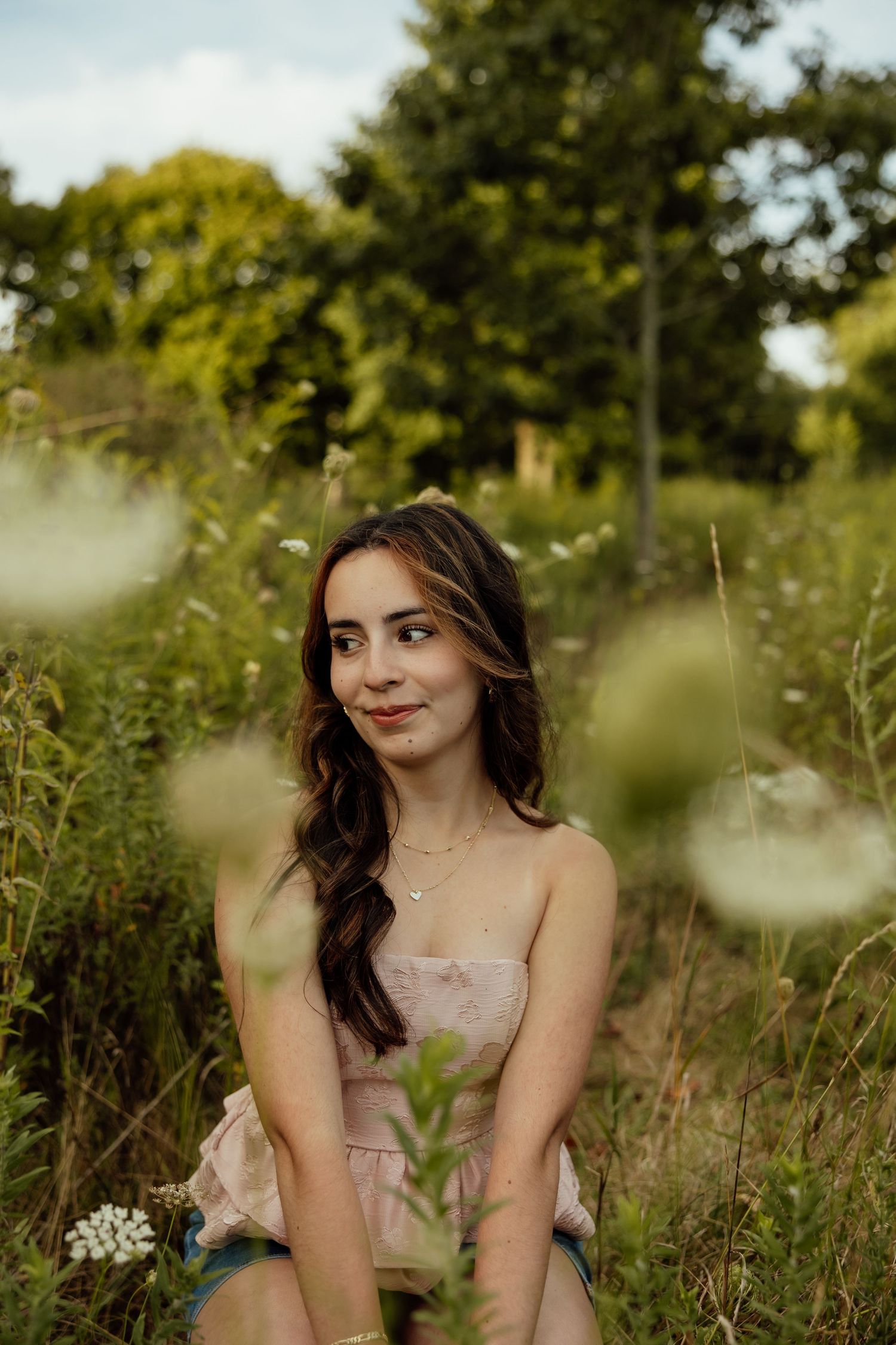 A person in a light colored dress poses gracefully in a sunlit meadow surrounded by wildflowers and greenery.