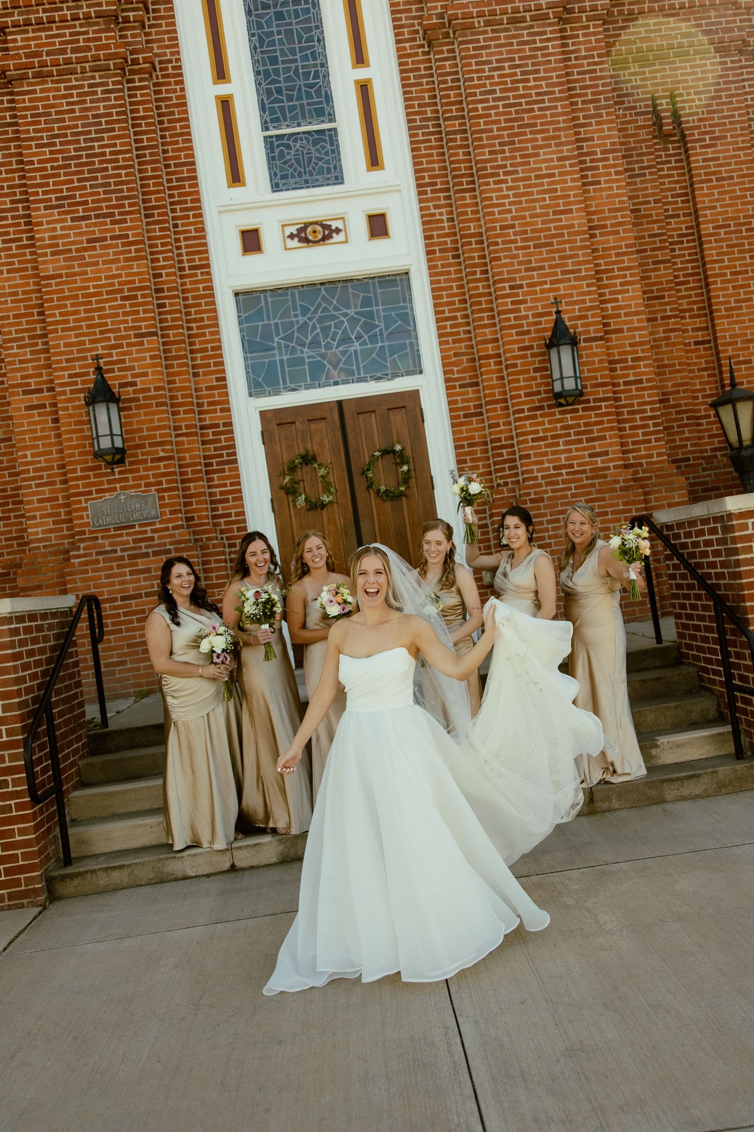 Wedding party in beige dresses posing outside brick church with stained glass windows.