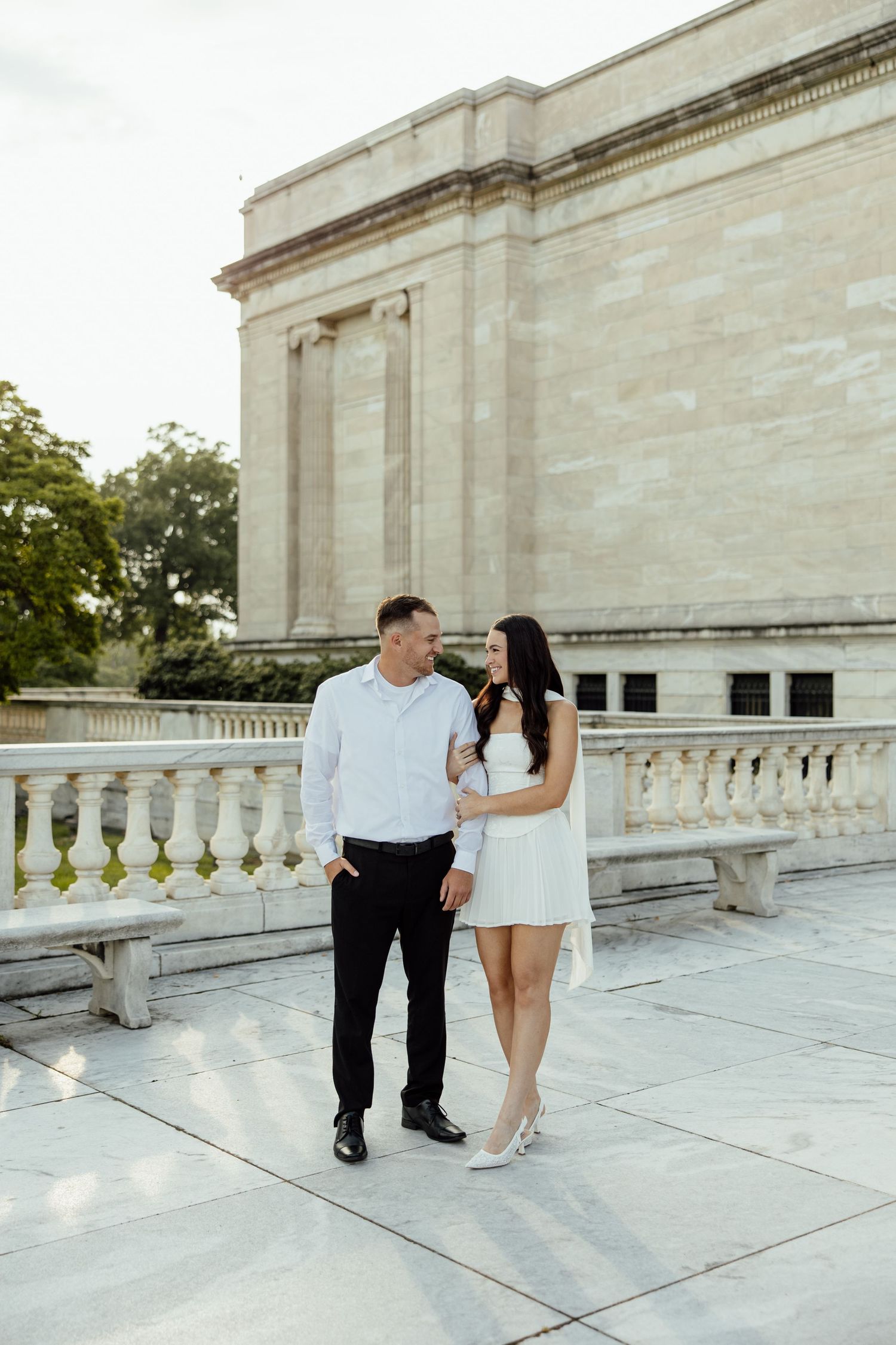 A couple in white and black outfits share romantic moments on a light stone balcony of a classical building.