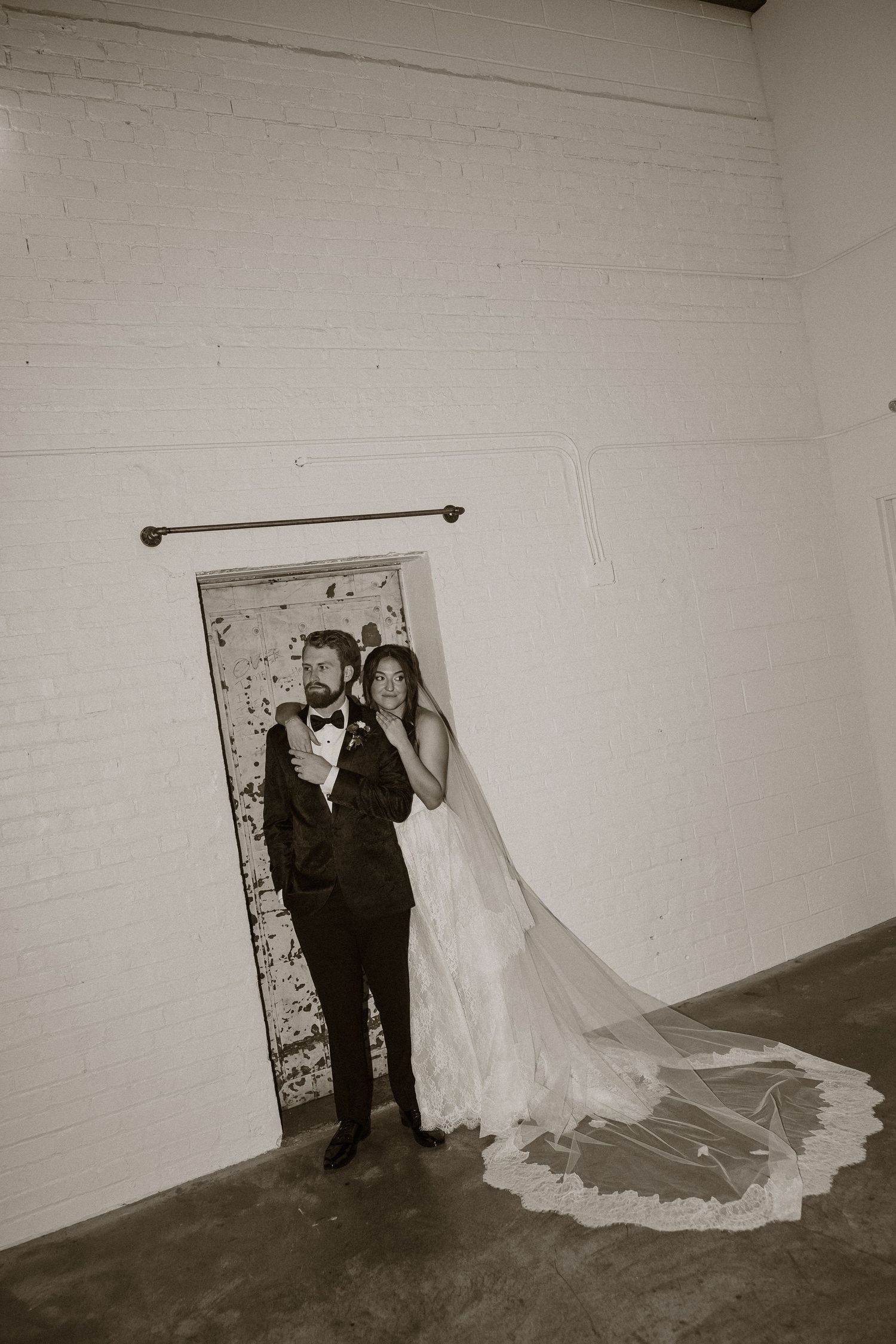 Black and white wedding portrait series against a white brick wall showing a flowing dress train.