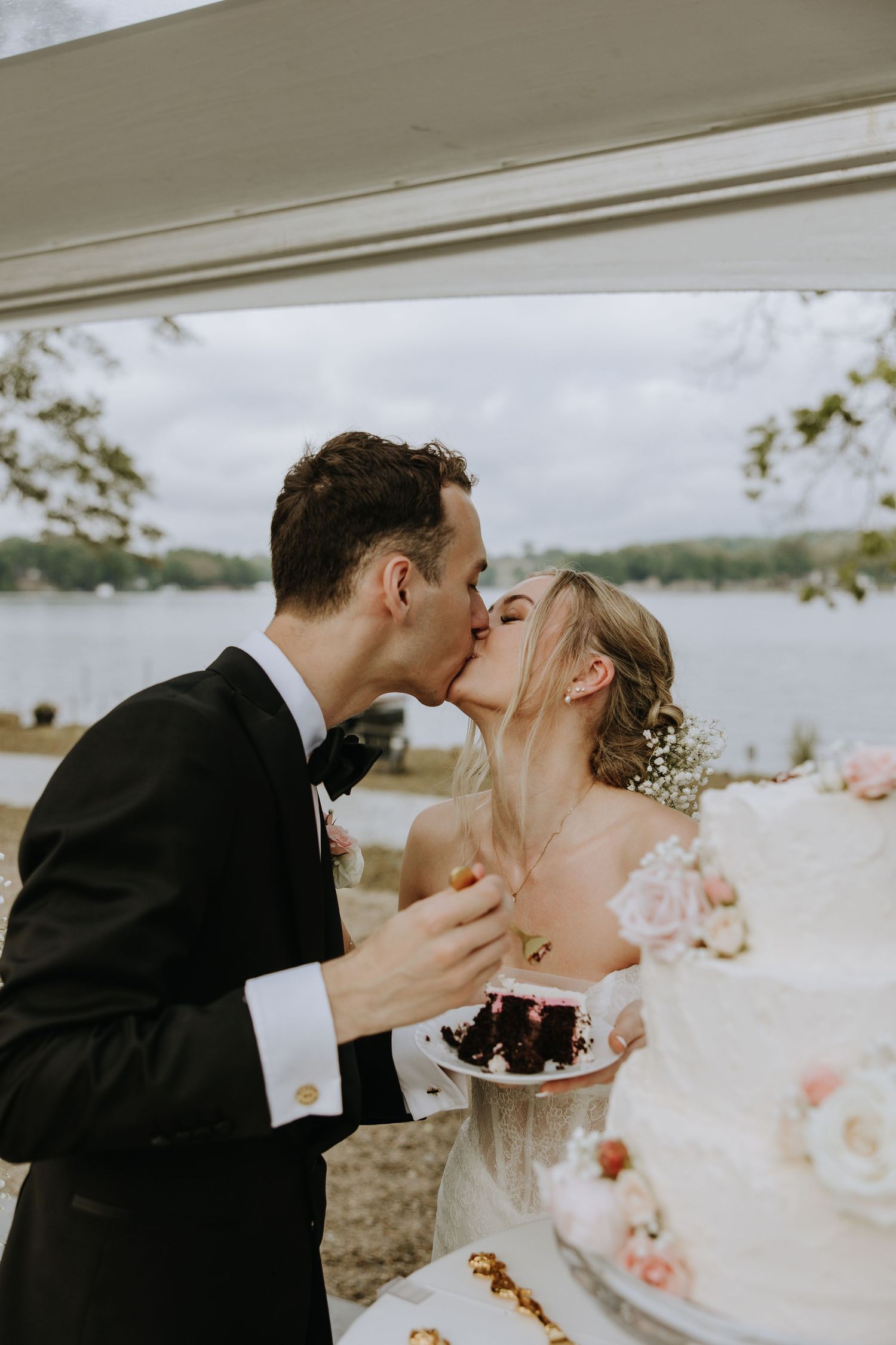 Newlyweds sharing cake by lakeside with delicate floral decorations.