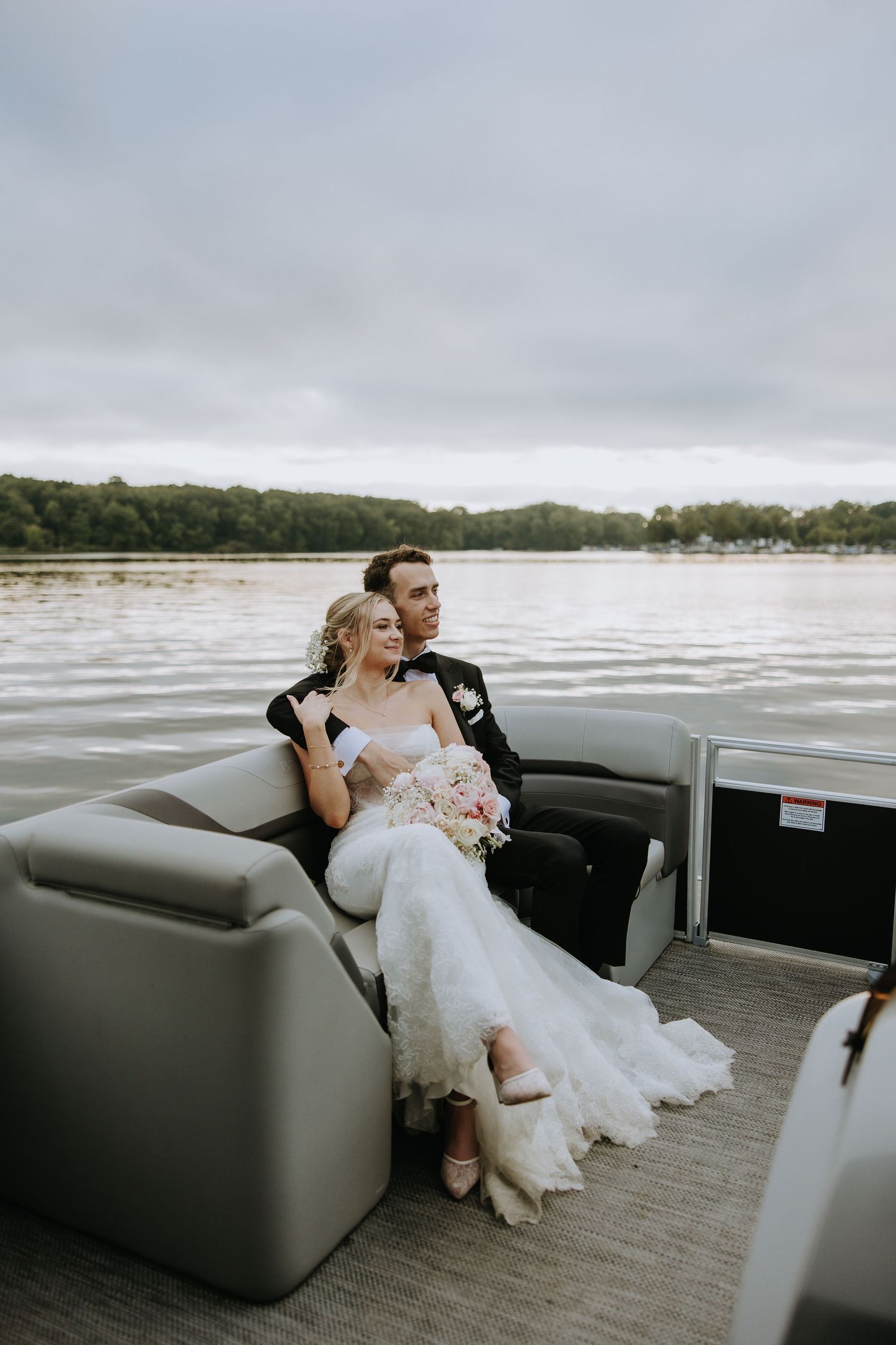 A couple relaxes together on a pontoon boat bench seat while cruising along a scenic waterfront on their wedding day.