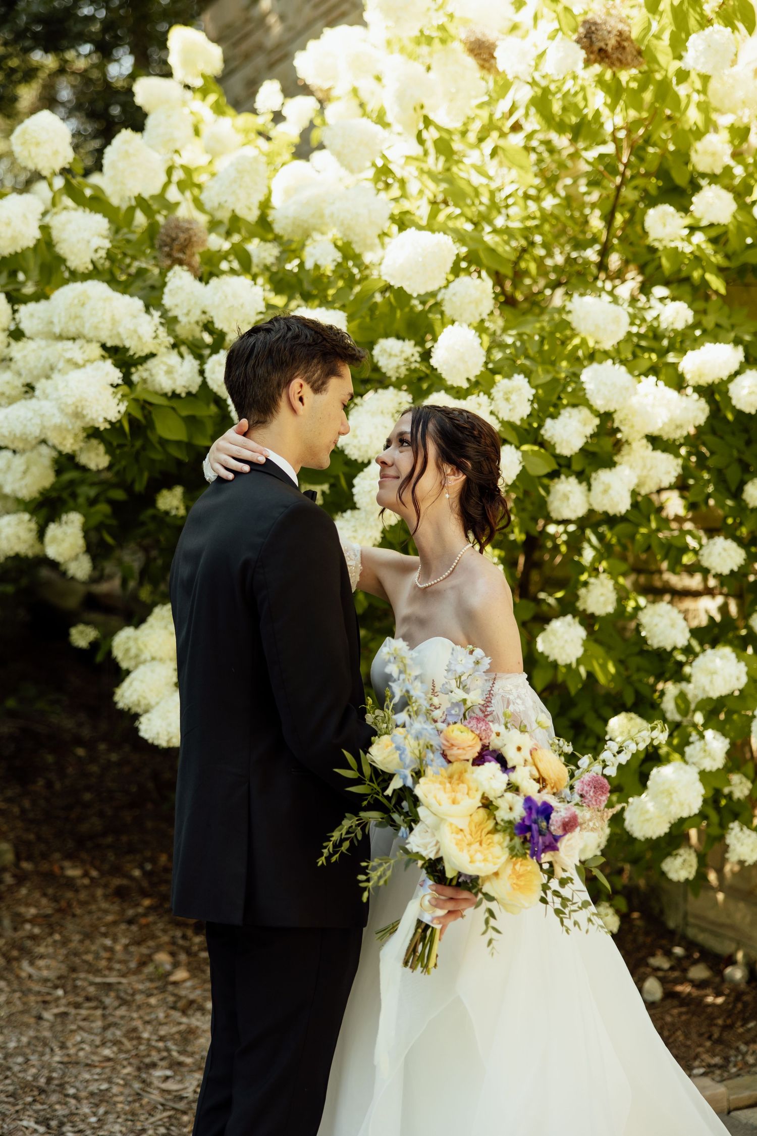 A romantic wedding moment captured in front of blooming white hydrangea bushes with a colorful bouquet.