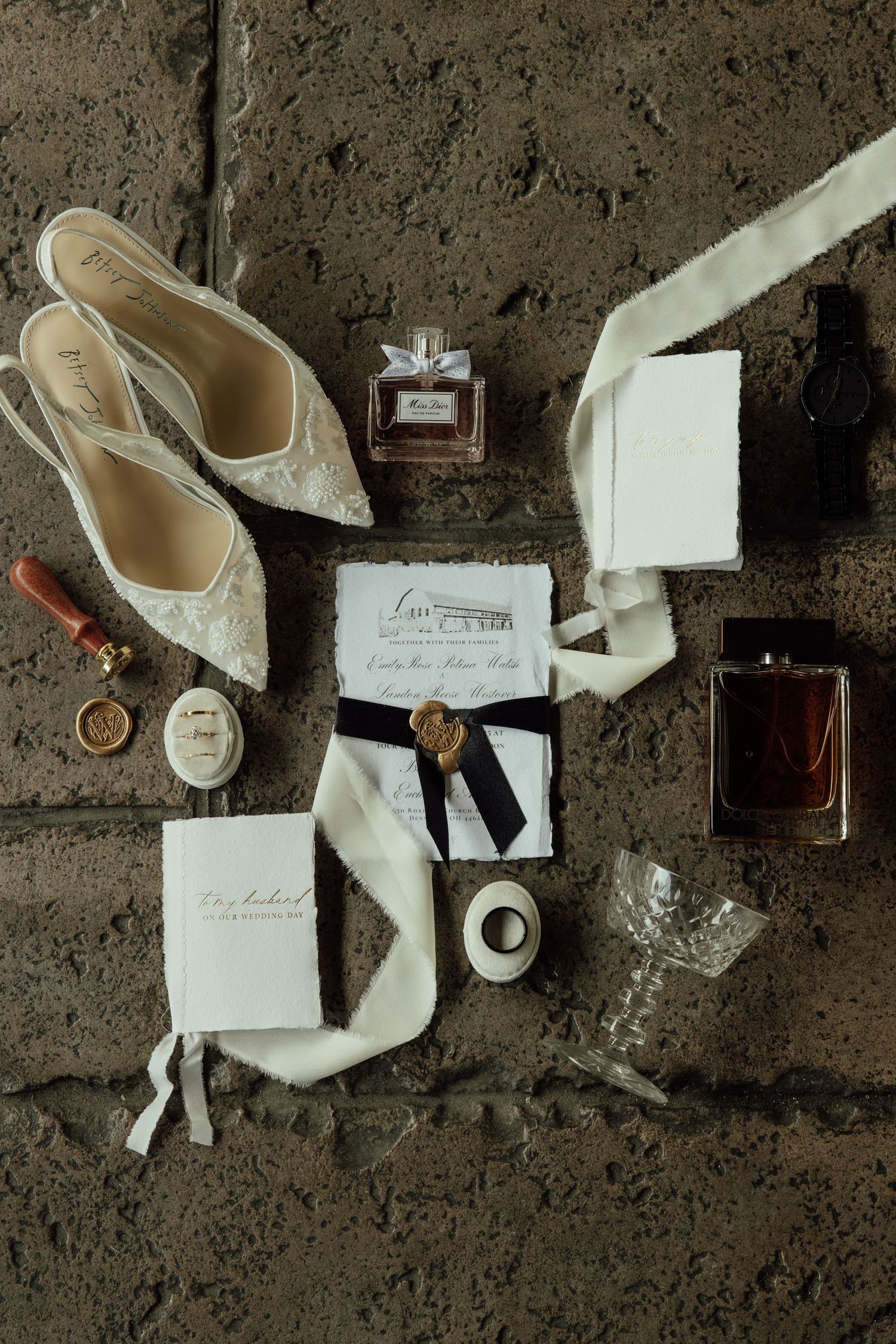 Elegant wedding accessories including lace heels, perfume bottles, invitations and ribbons arranged on concrete surface.