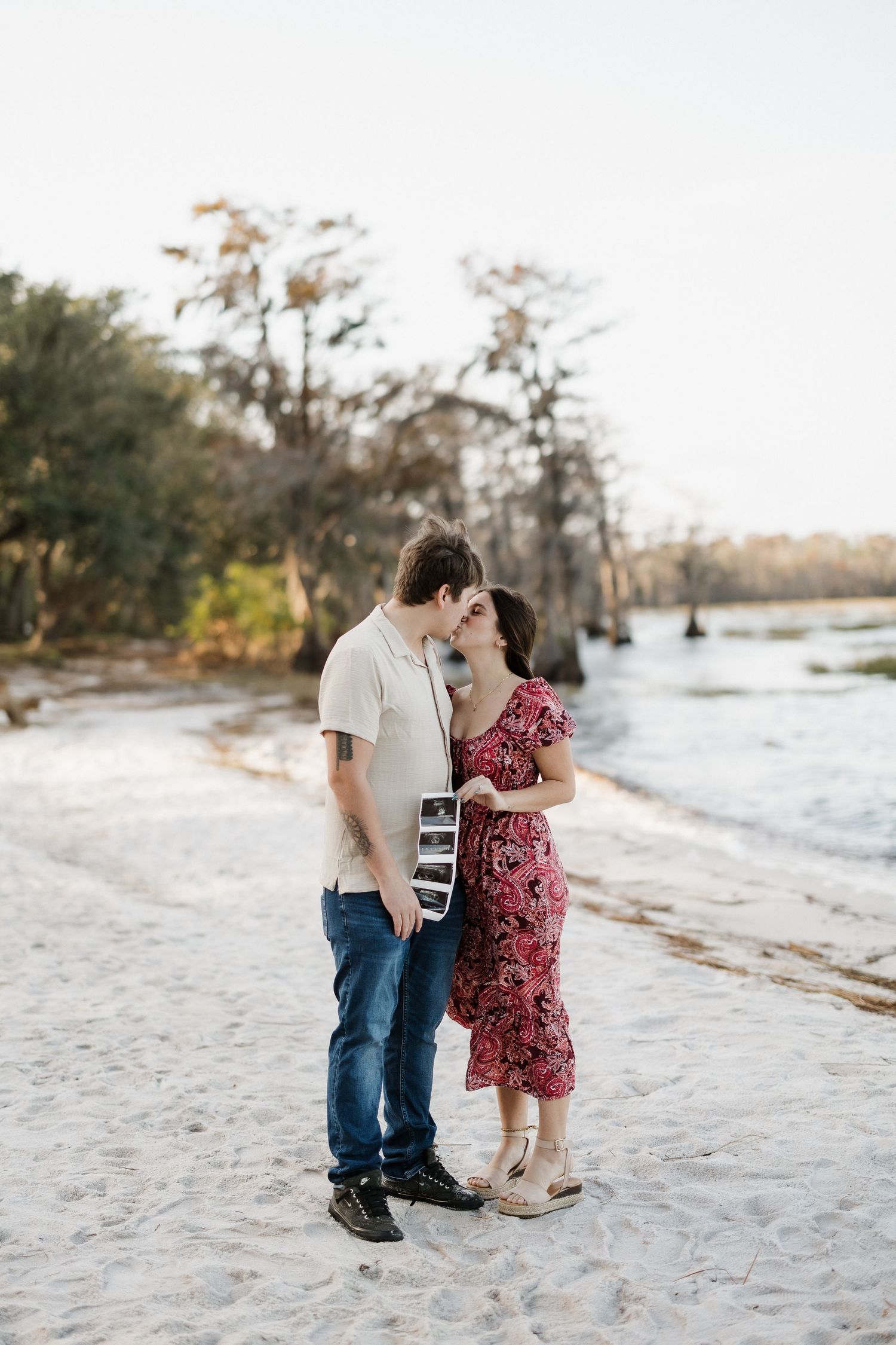 Couple sharing tender moment on beach while holding ultrasound photos.