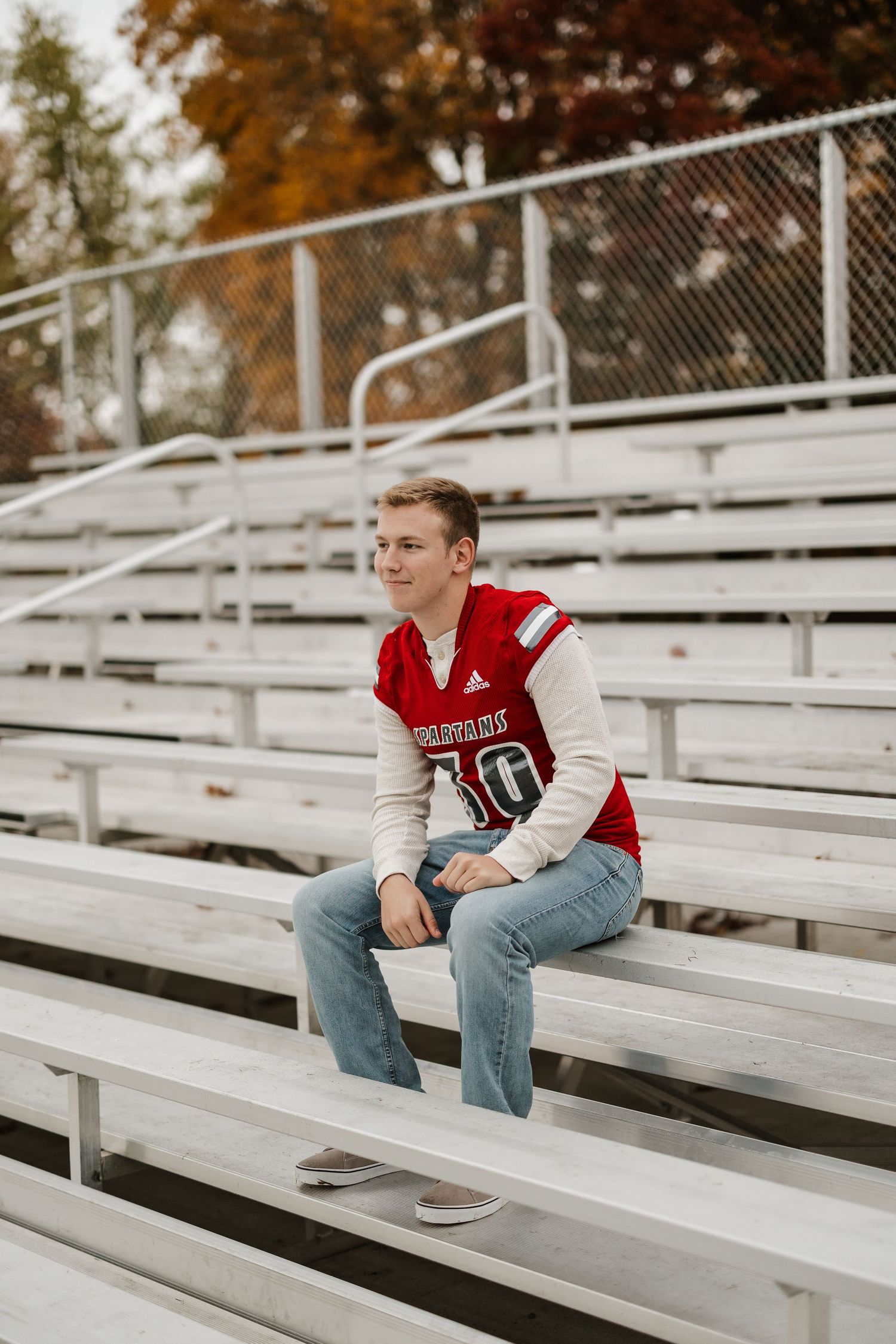 Student in varsity jacket sits thoughtfully on bleachers during autumn season.