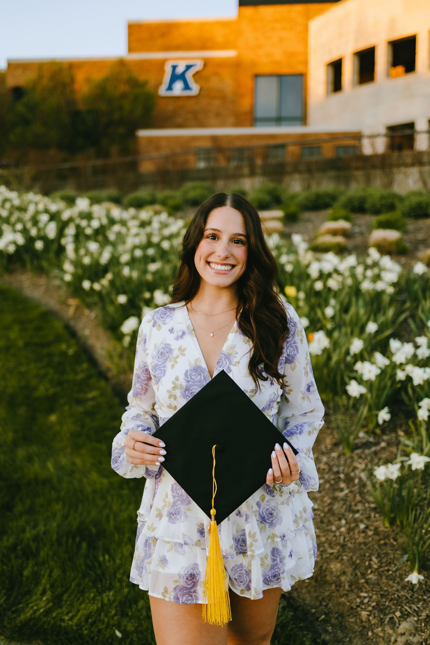 A graduate holds their black cap with yellow tassel while standing in front of a university building.