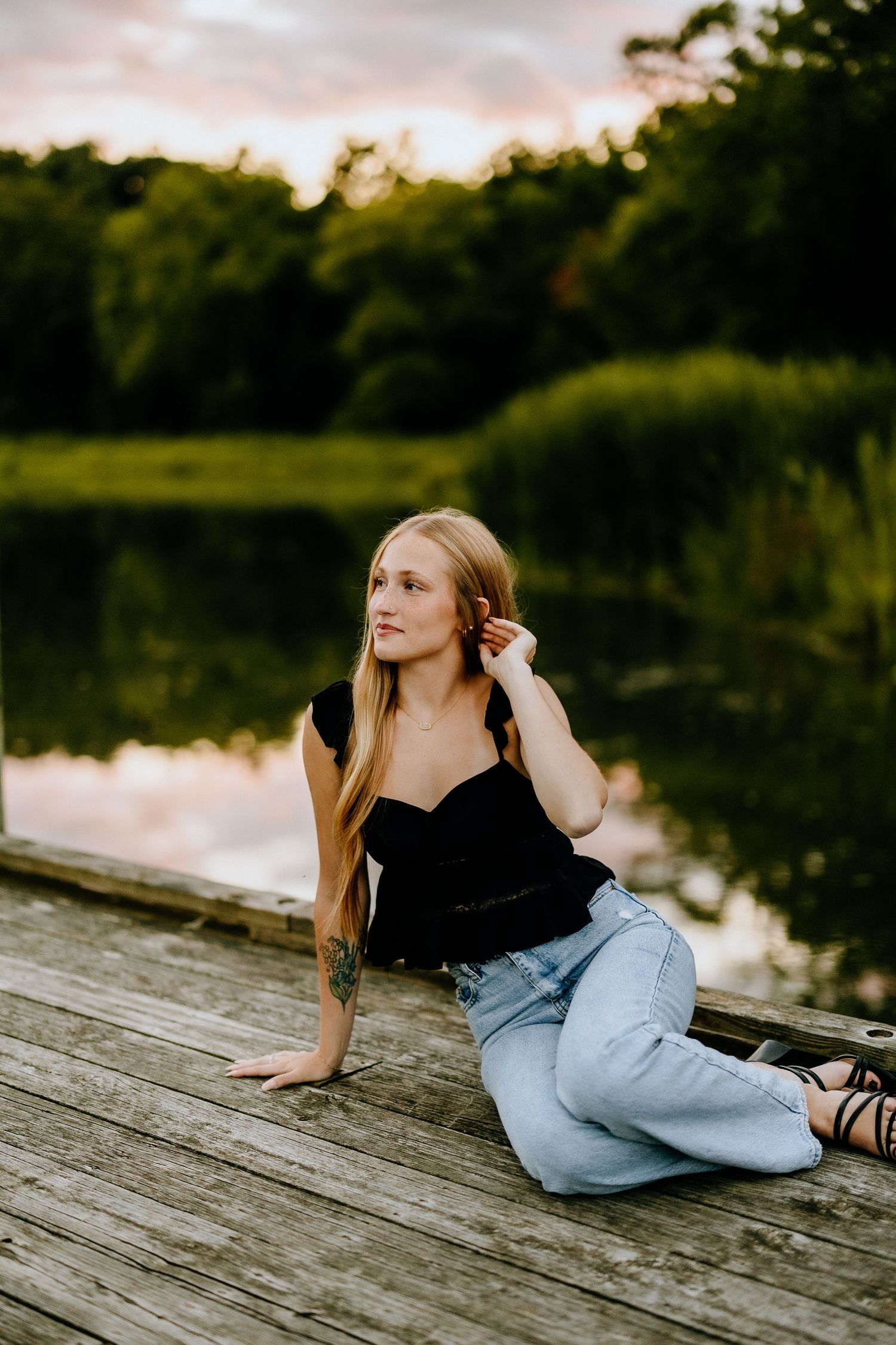 Person in black top and jeans poses on wooden dock by lake during sunset.