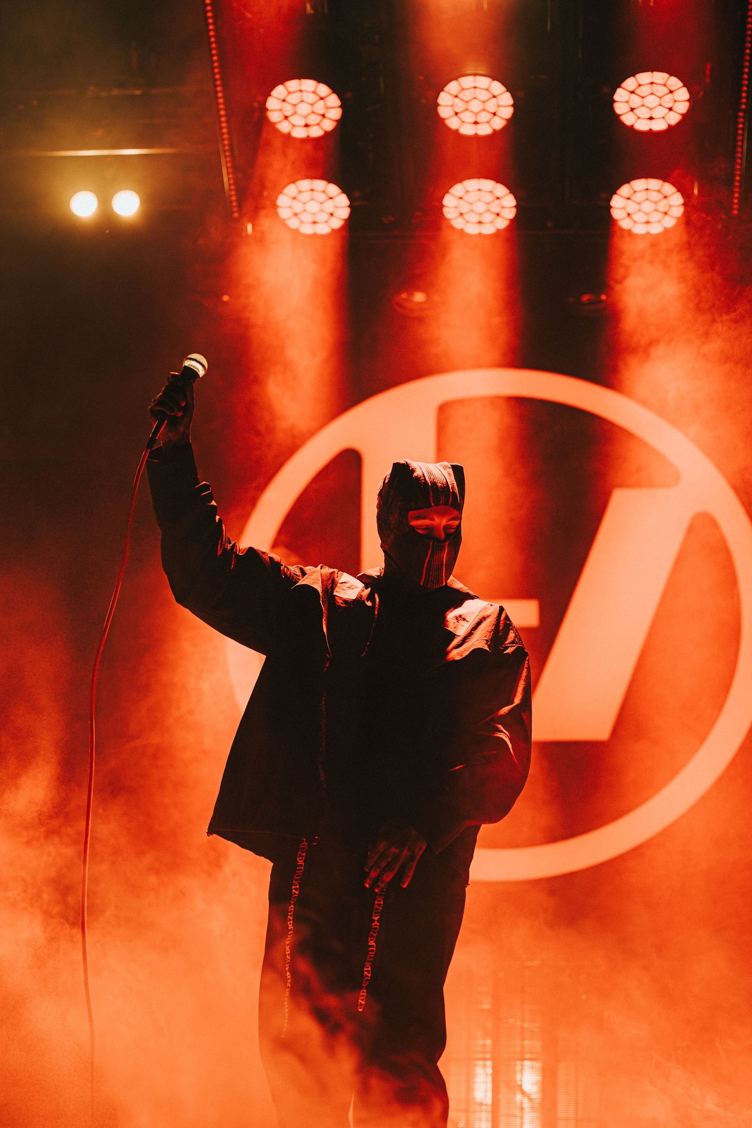 A dramatic concert performance silhouette against red stage lighting and LED spotlights with a circular logo backdrop.