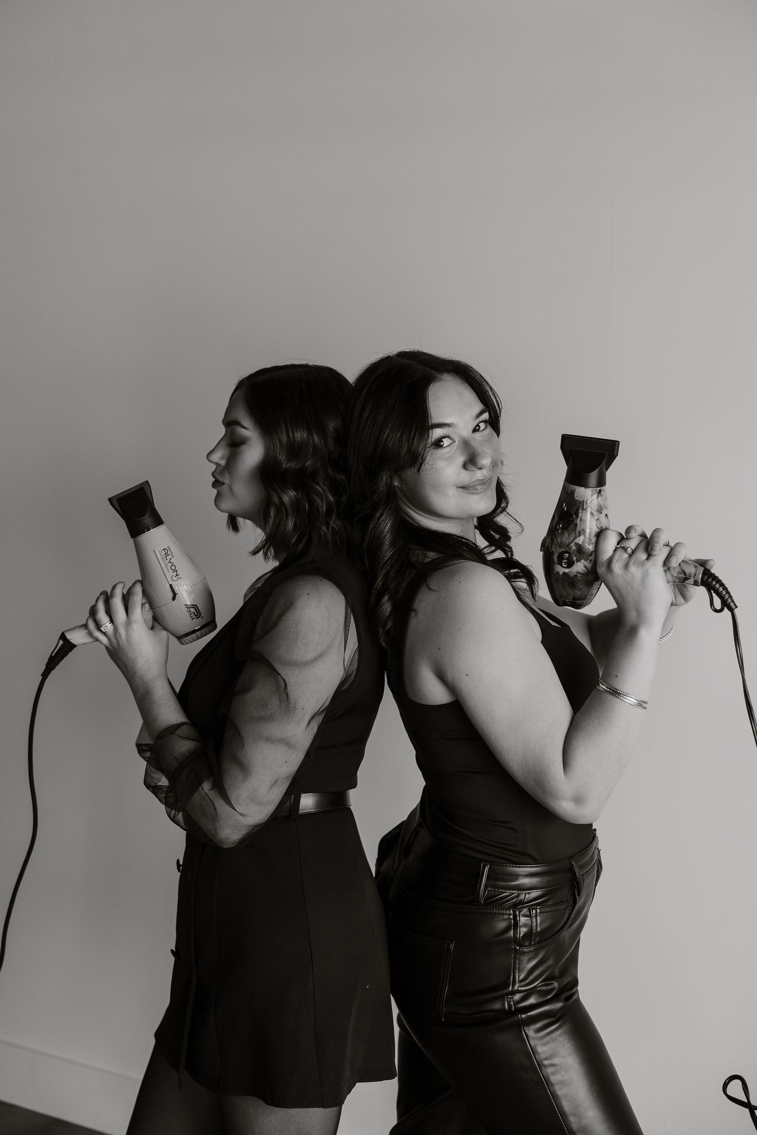 Black and white portrait of two hairstylists back-to-back holding styling tools.