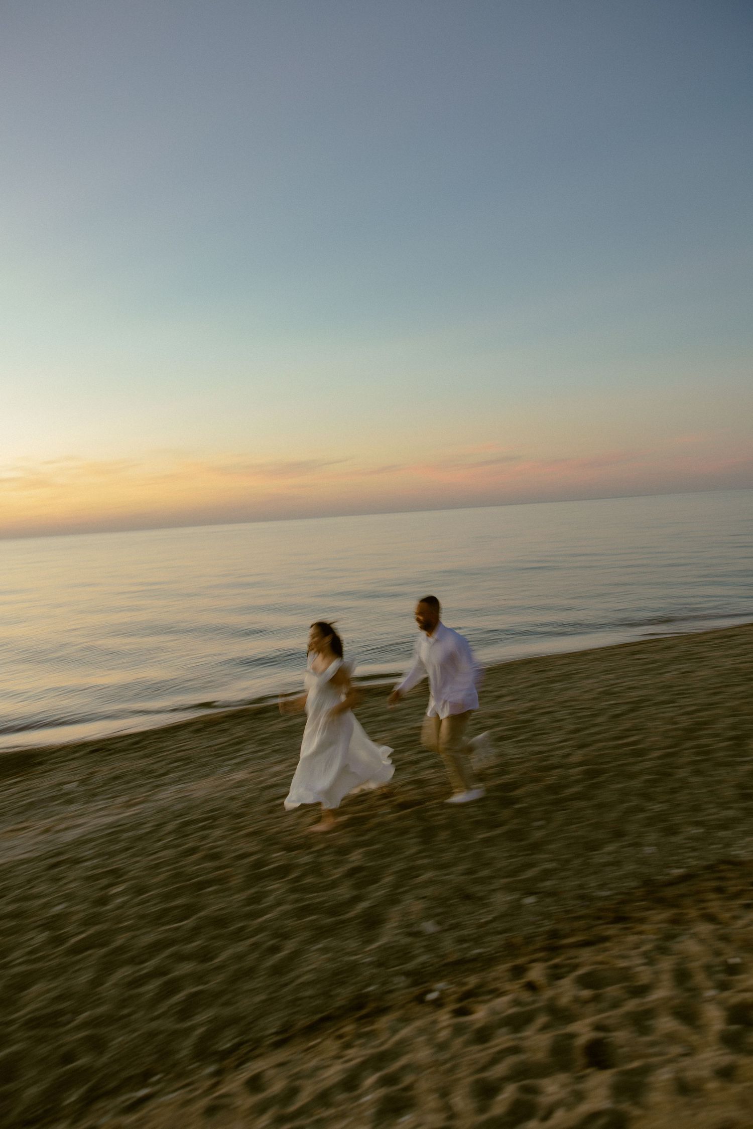 Two people in white outfits walk along a beach at sunset with golden light casting across the ocean horizon.