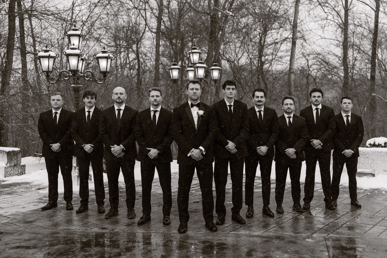 A row of groomsmen in black suits stand in line on a snowy winter day for a formal wedding photo.