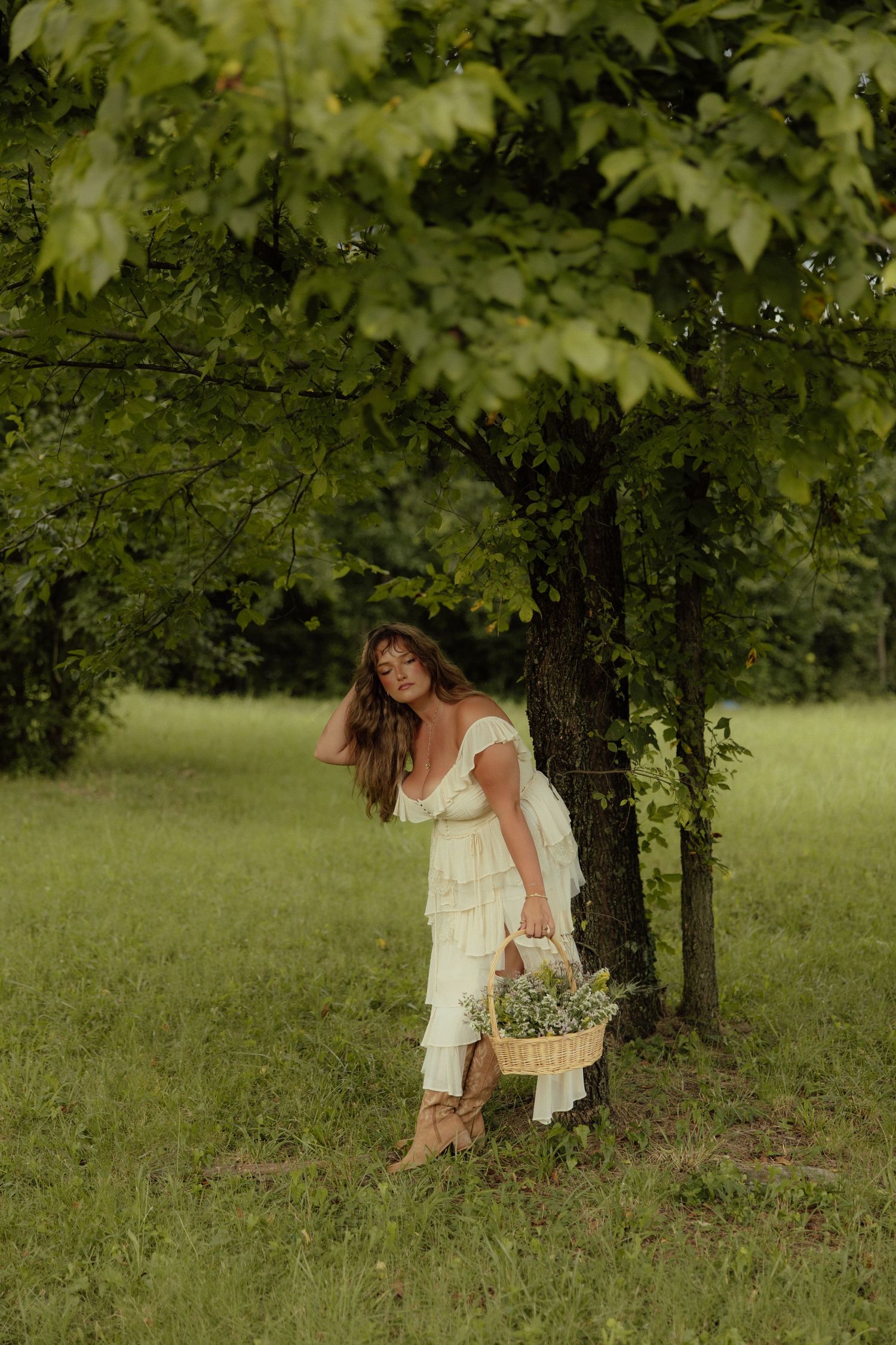 A figure in a white dress gathers wildflowers in a lush green meadow beneath shady trees.