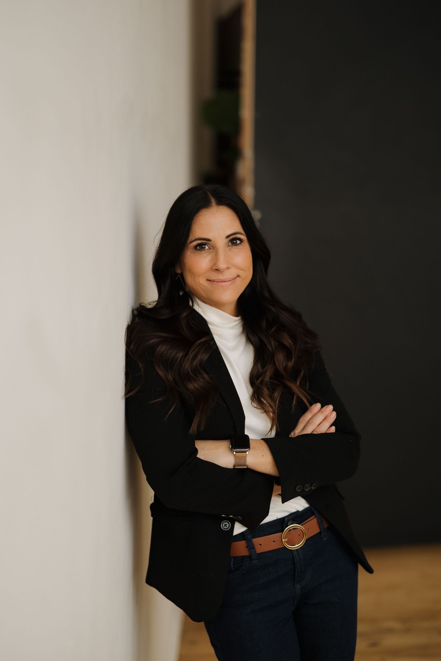 Professional standing pose in black blazer and white turtleneck against office wall.