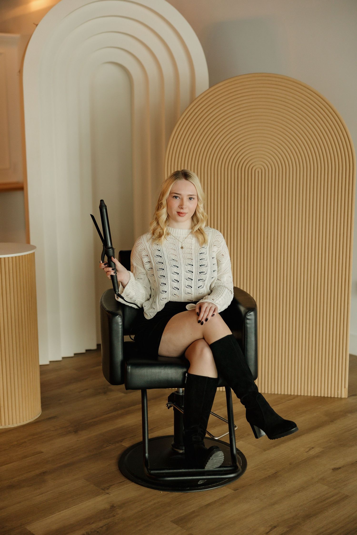 Seated portrait in a white sweater and boots against an arched wall with modern decor.