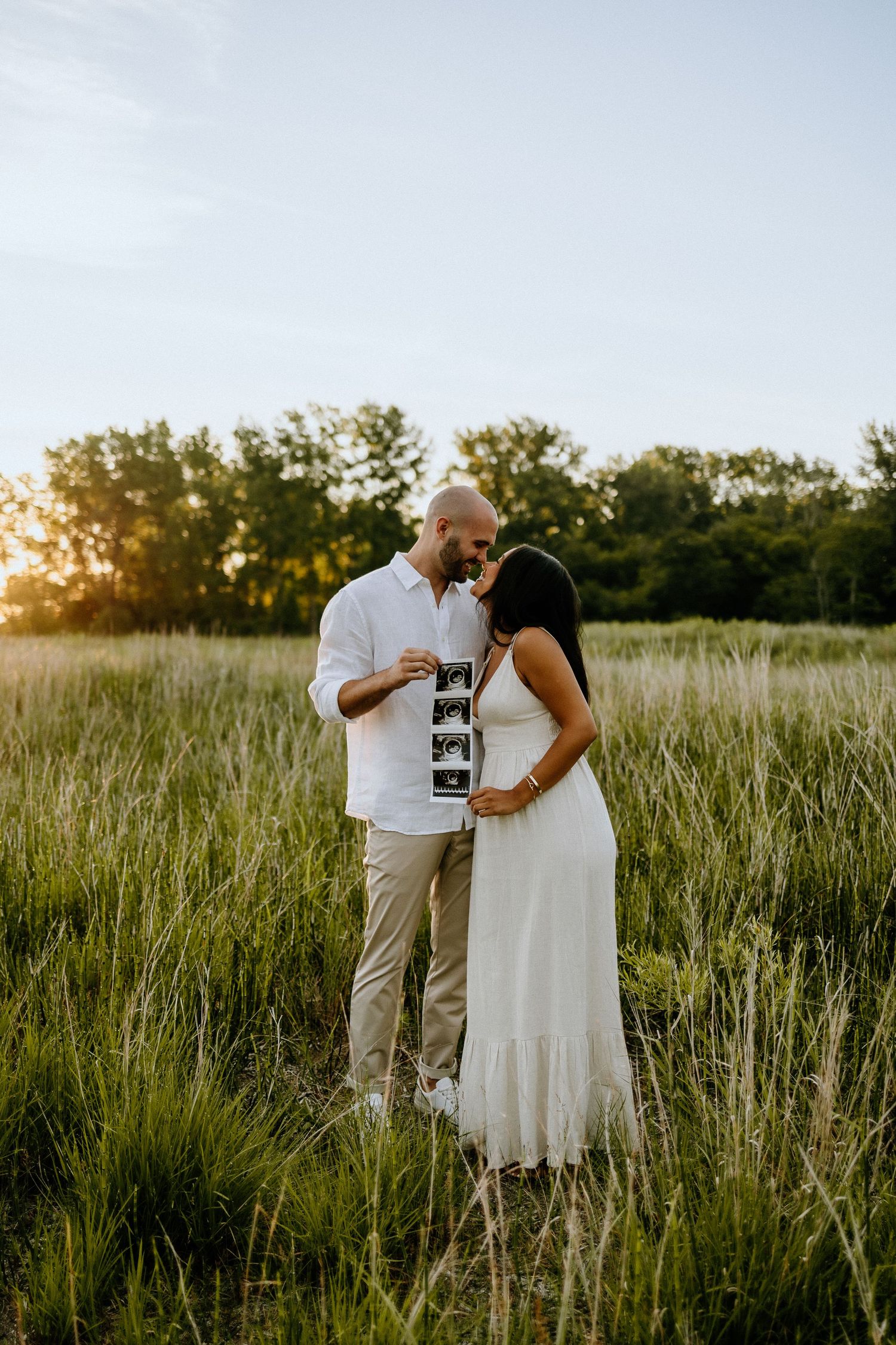 Couple holding ultrasound photos while standing in a sunlit meadow.