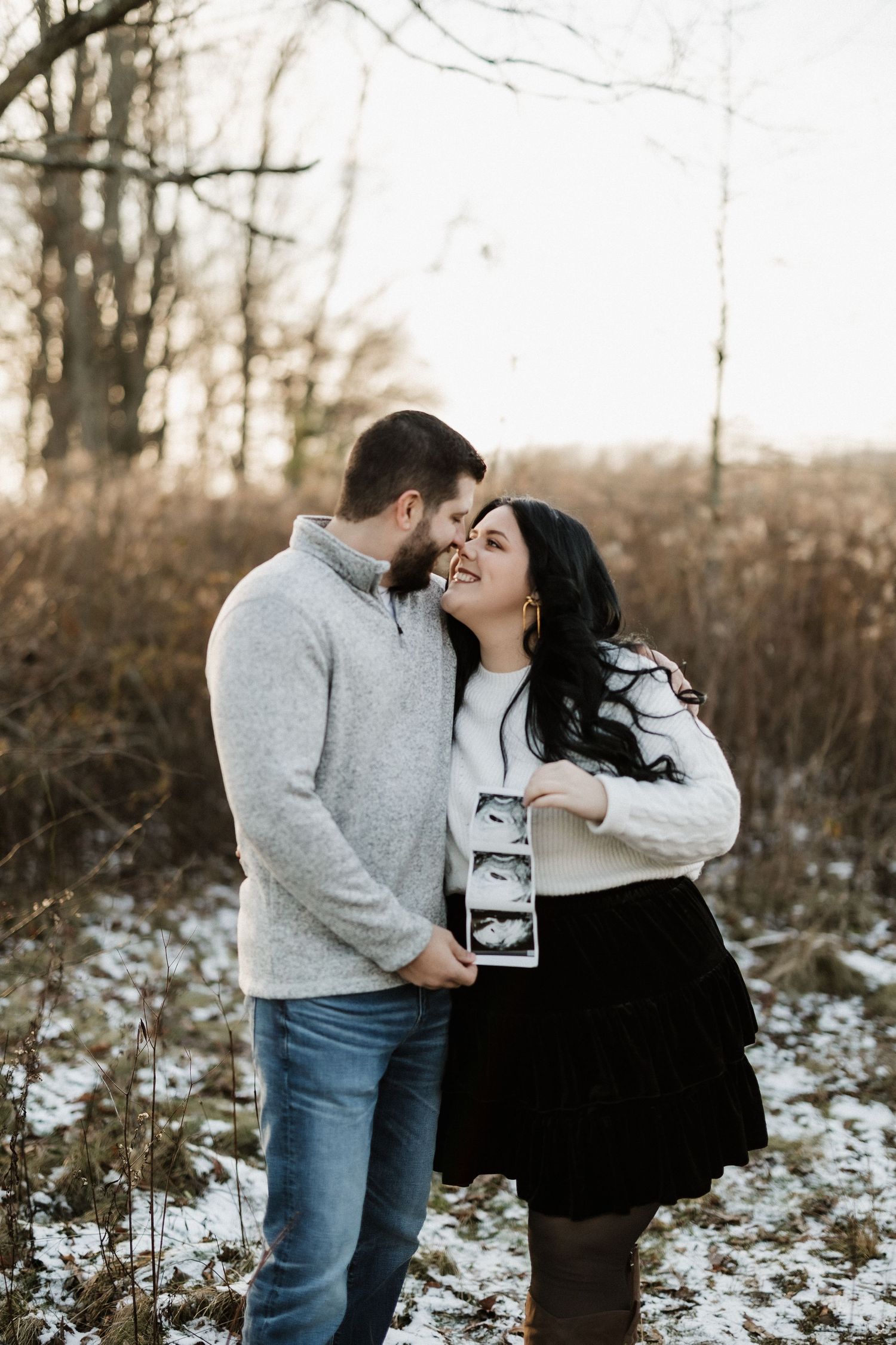 A winter pregnancy announcement photo with ultrasound images in a snowy outdoor setting.