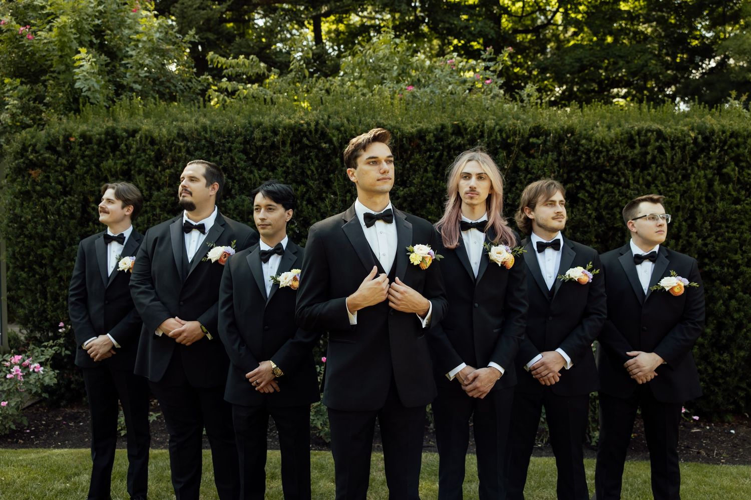 Groomsmen in black tuxedos lined up for formal wedding photos in garden setting.