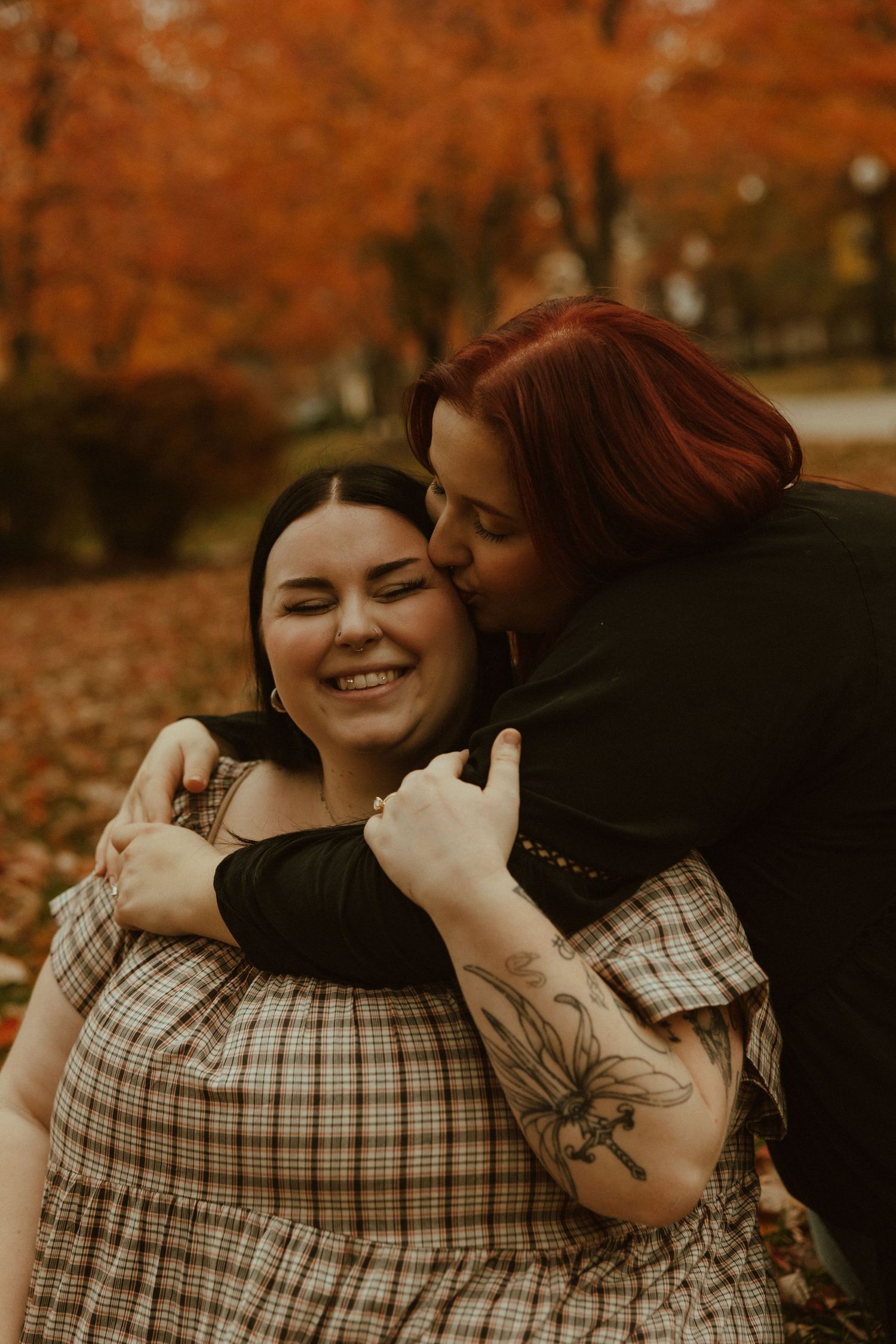 Two people share an intimate embrace while laughing together during an autumn photoshoot with orange foliage in background.