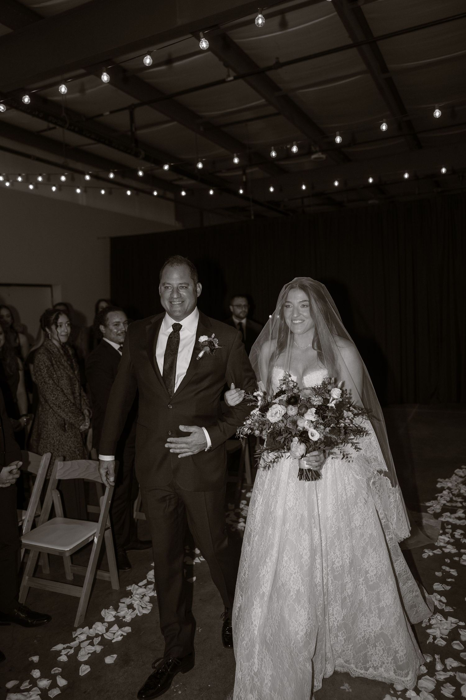 Wedding ceremony processional with string lights overhead and floral aisle decorations.