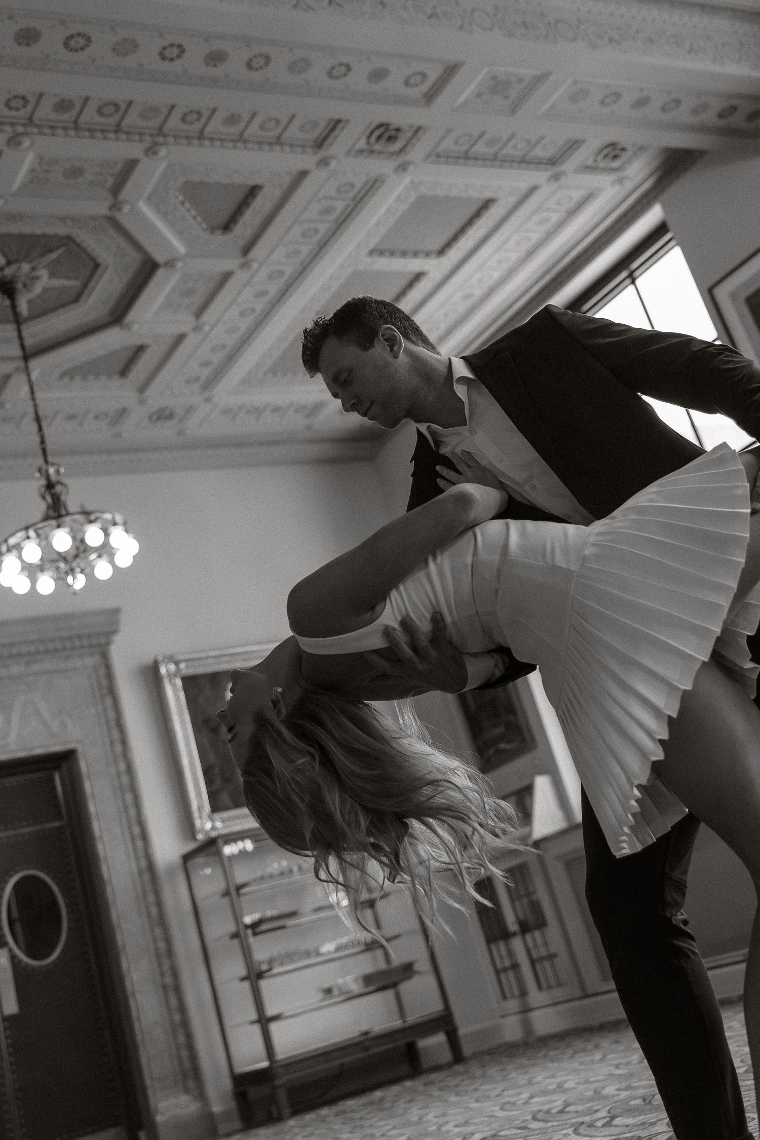 A black and white photograph of two dancers performing an elegant dip in an ornate ballroom with a chandelier.