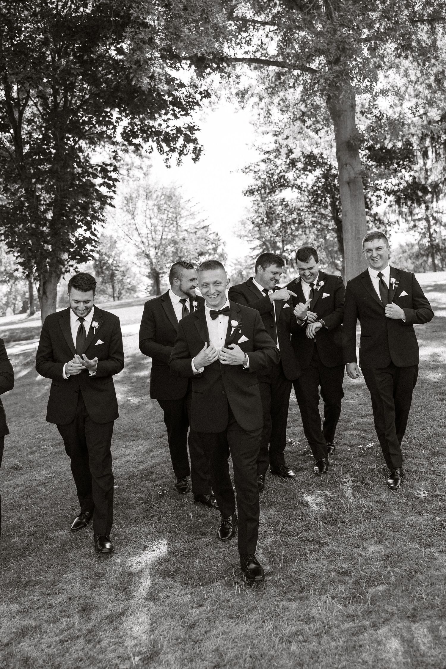 A black and white photo of groomsmen walking together outdoors on a lawn surrounded by trees.