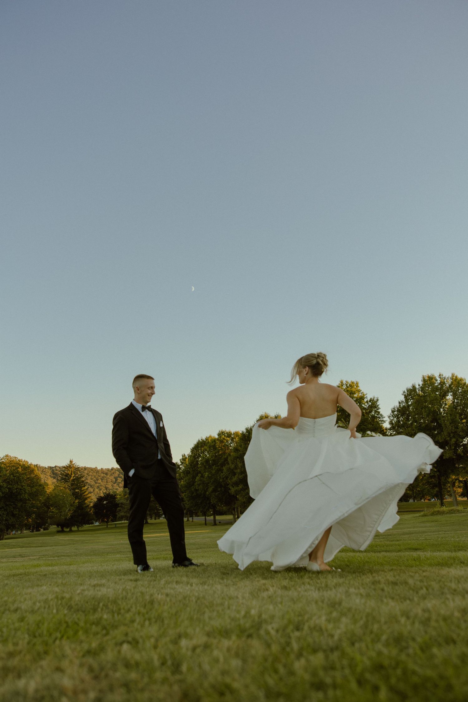 Newlyweds walking through a green field at sunset with flowing white dress.