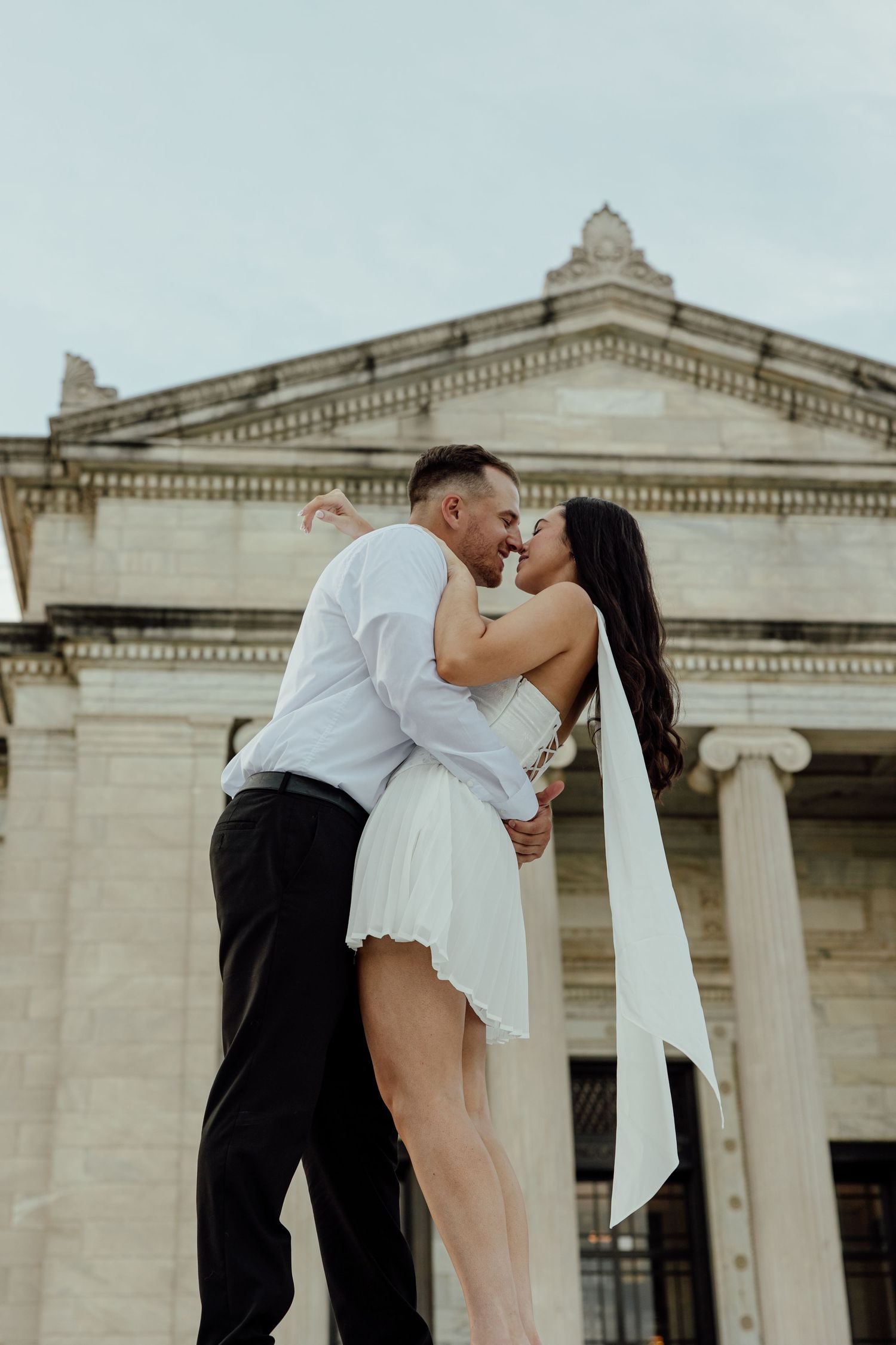 A romantic couple embraces in front of a grand neoclassical building with ornate columns and architecture.