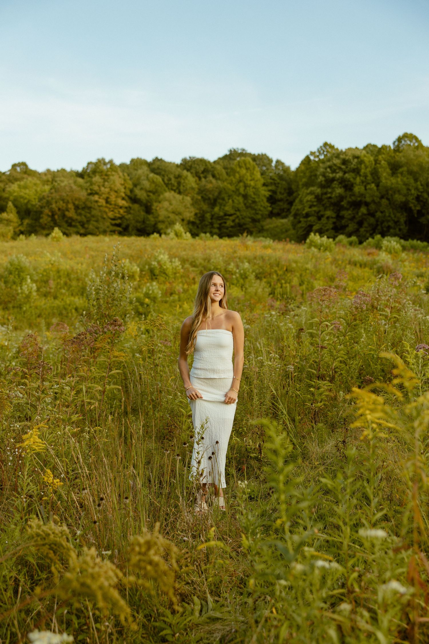 A person in a white dress stands in a golden meadow surrounded by tall wildflowers and greenery.