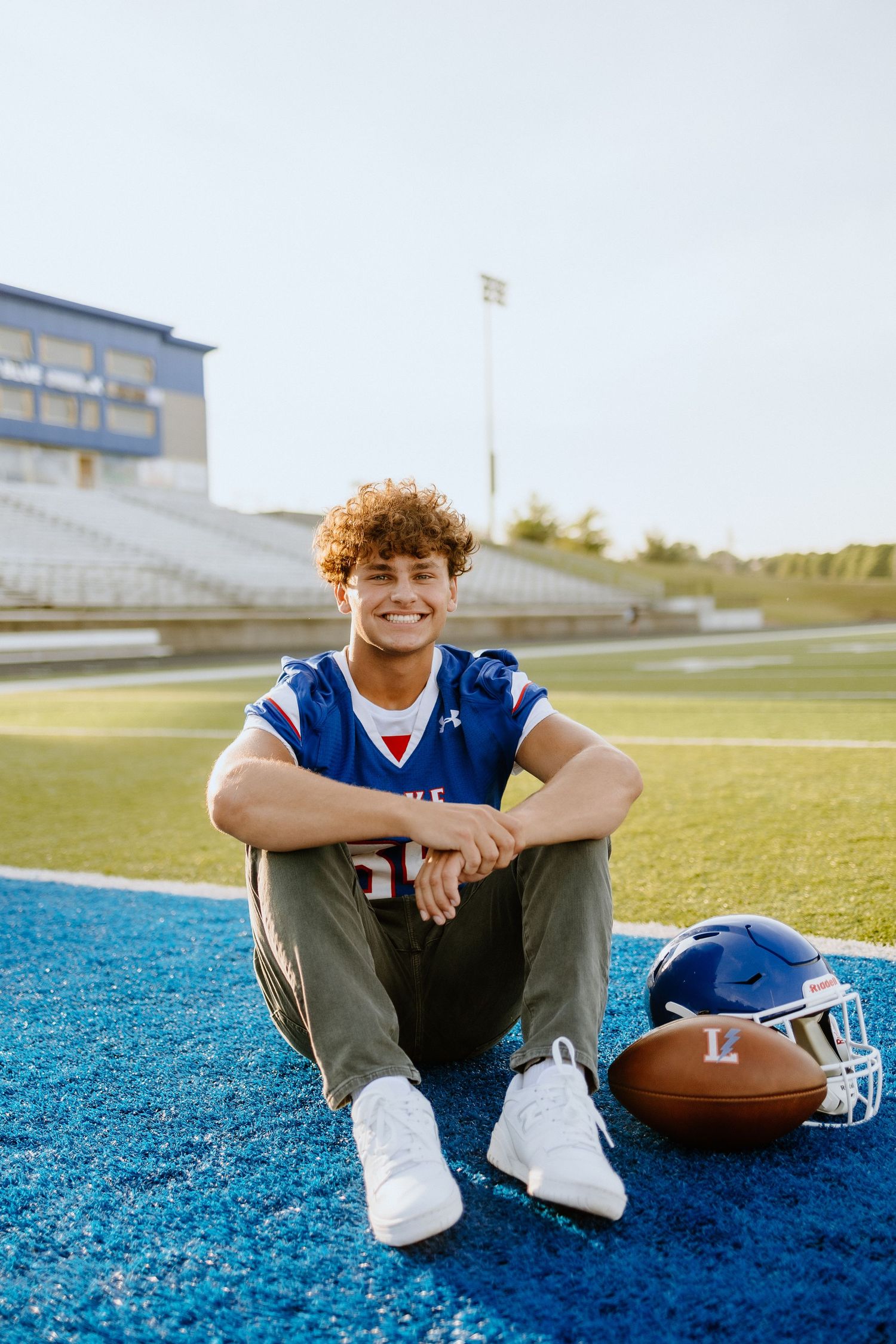 Athletic portrait on blue turf field with football and helmet at sunset.