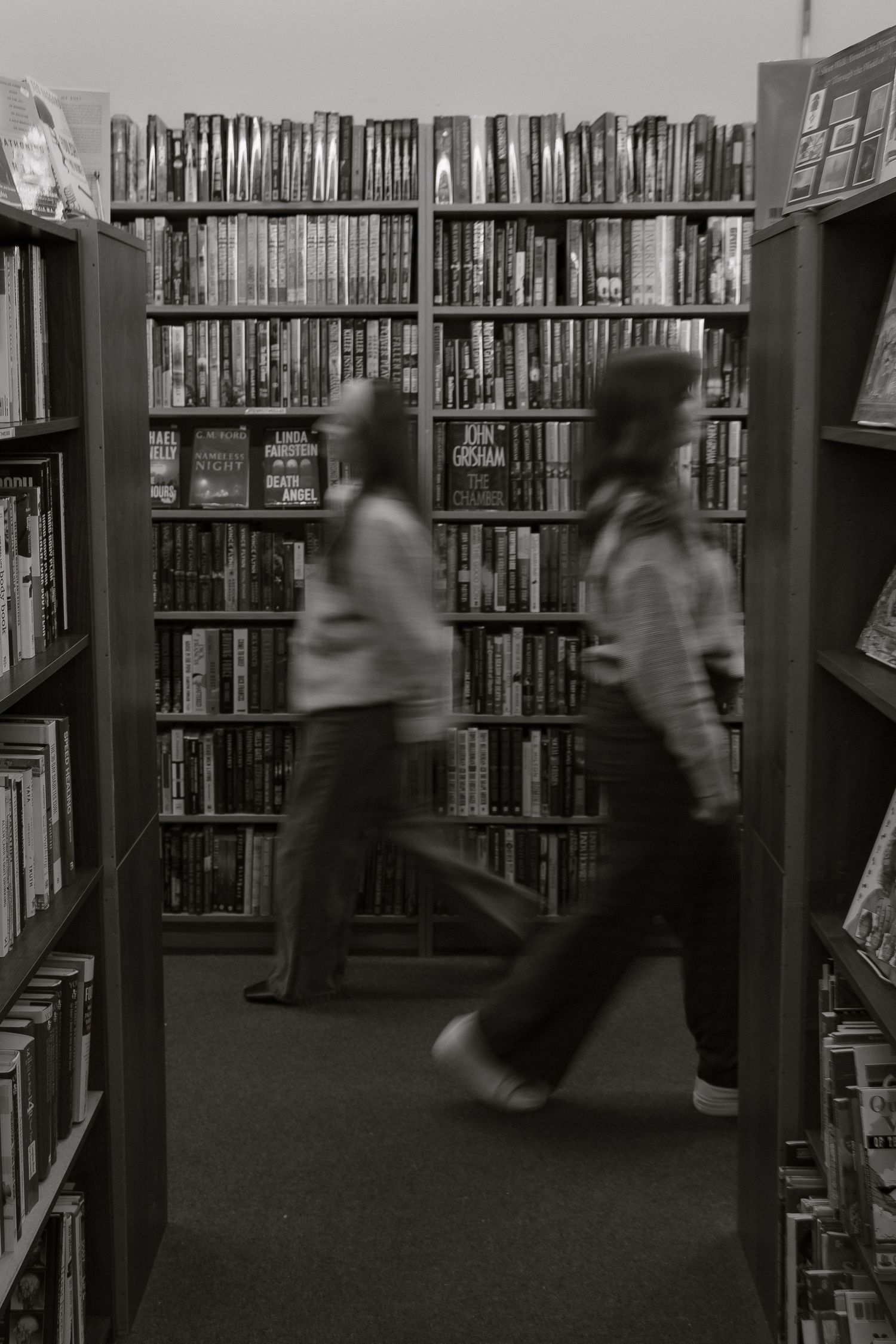 Blurred motion of people walking between tall library bookshelves in black and white.