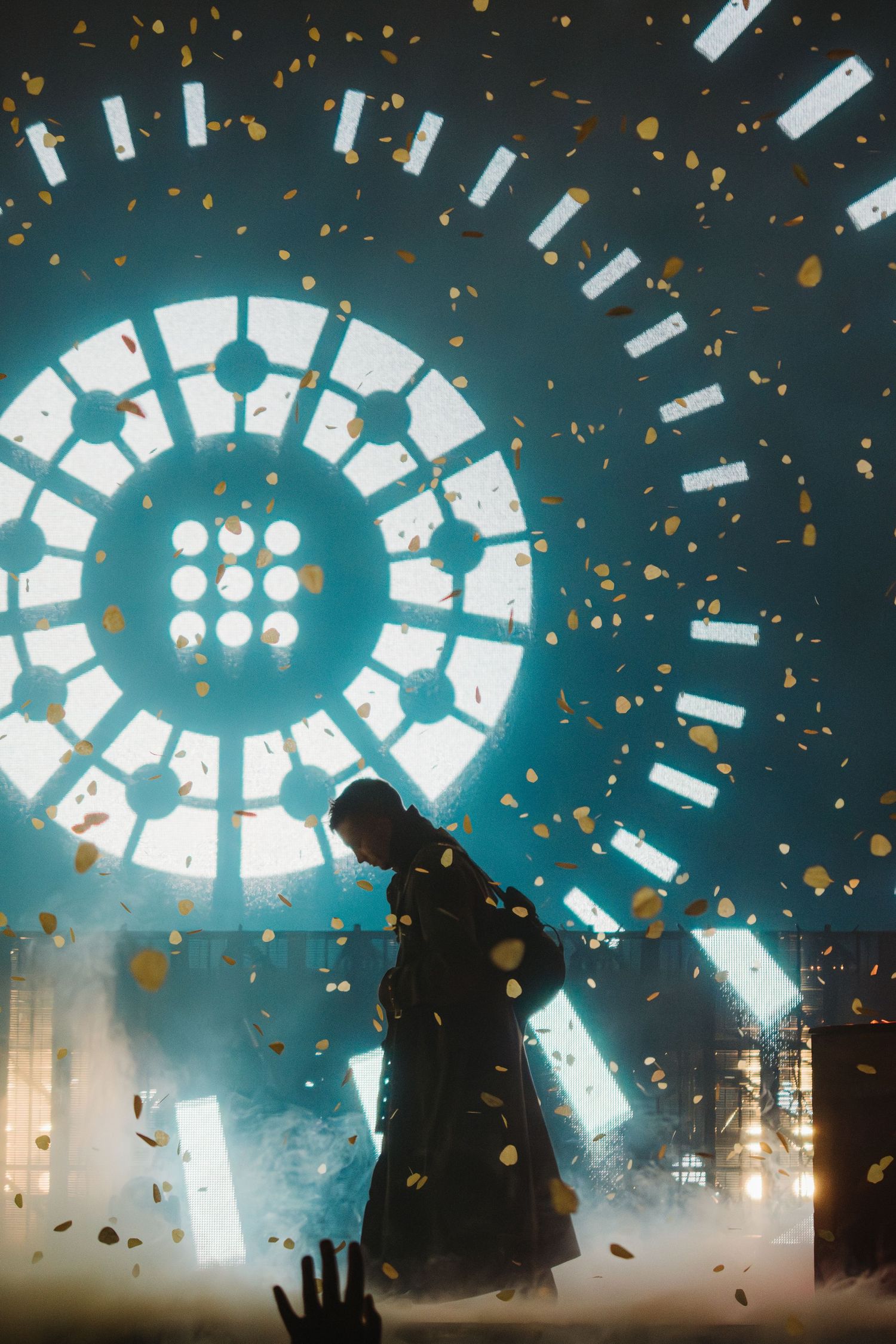 Performer silhouetted against circular light display with confetti falling during concert.
