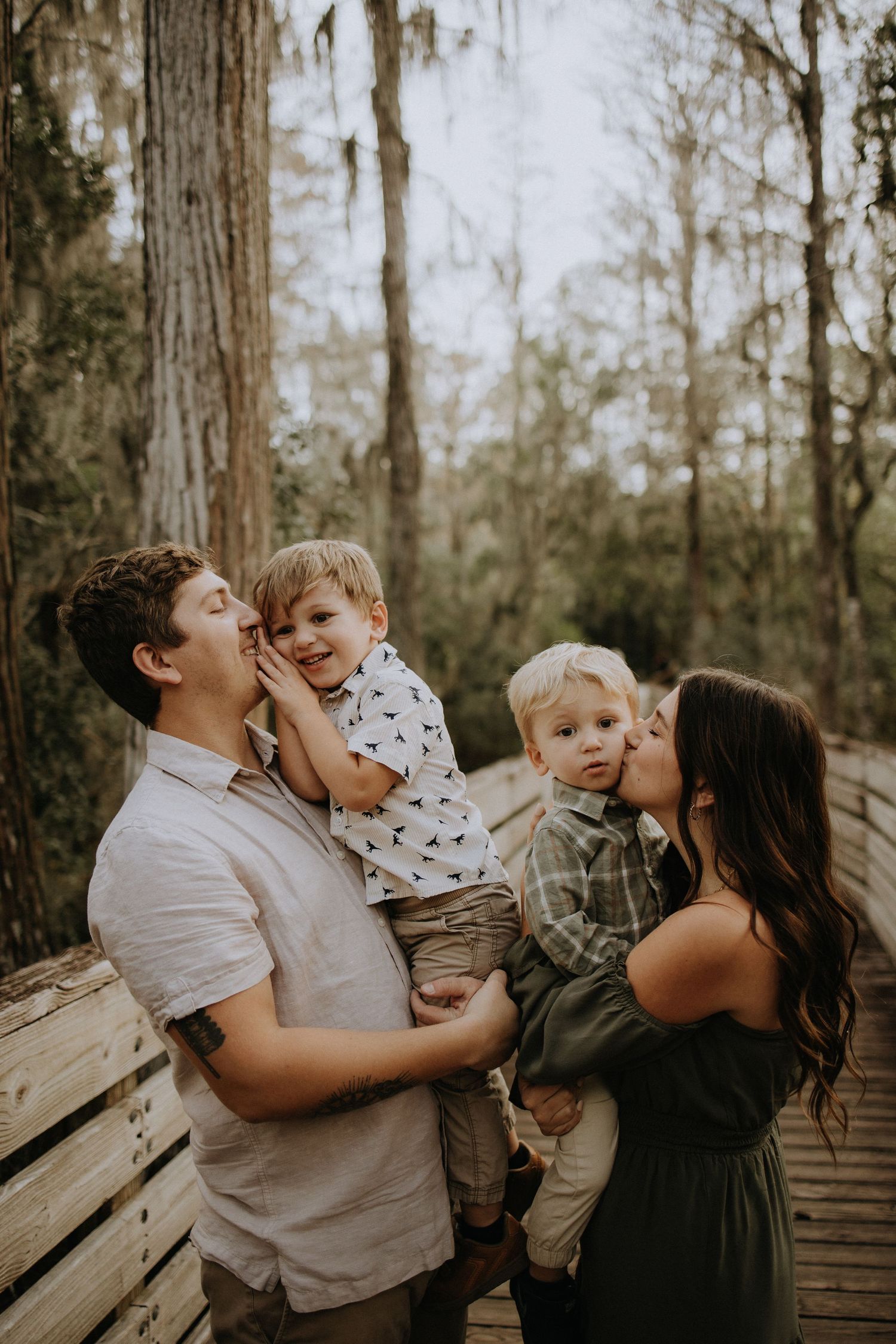 Family of four sharing intimate moments on wooden boardwalk in forest setting.