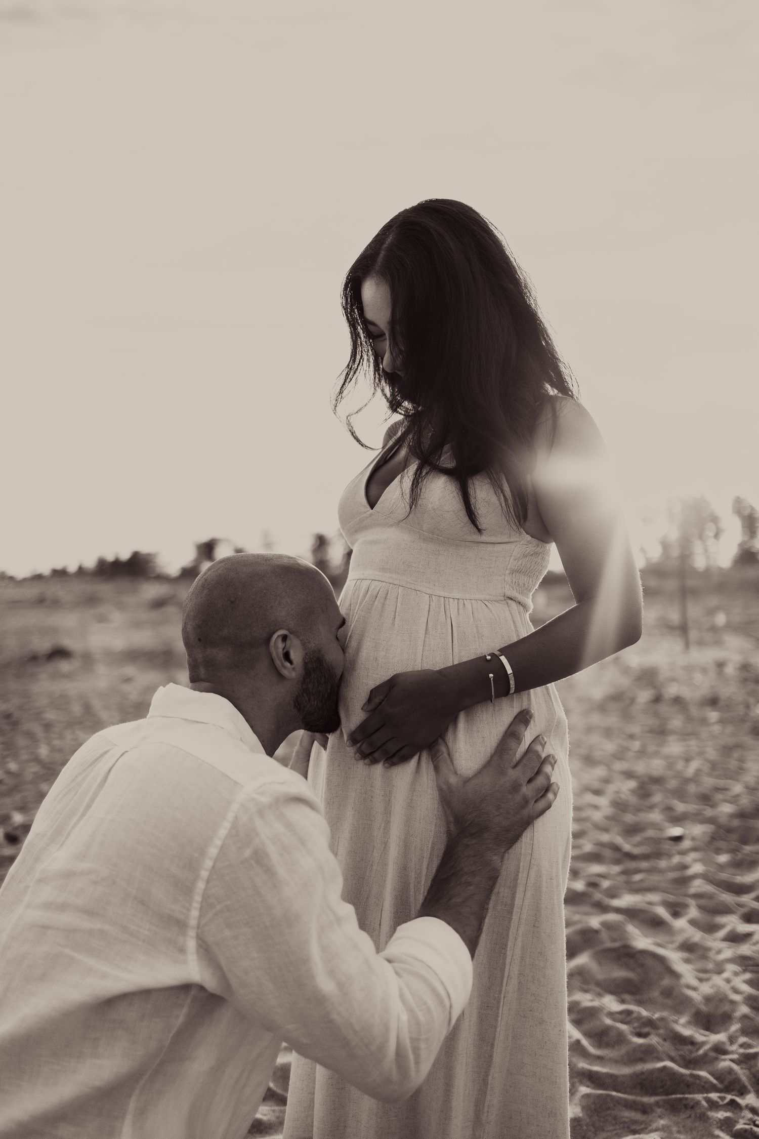 A tender maternity moment captured on a beach with a couple in white clothing at sunset.