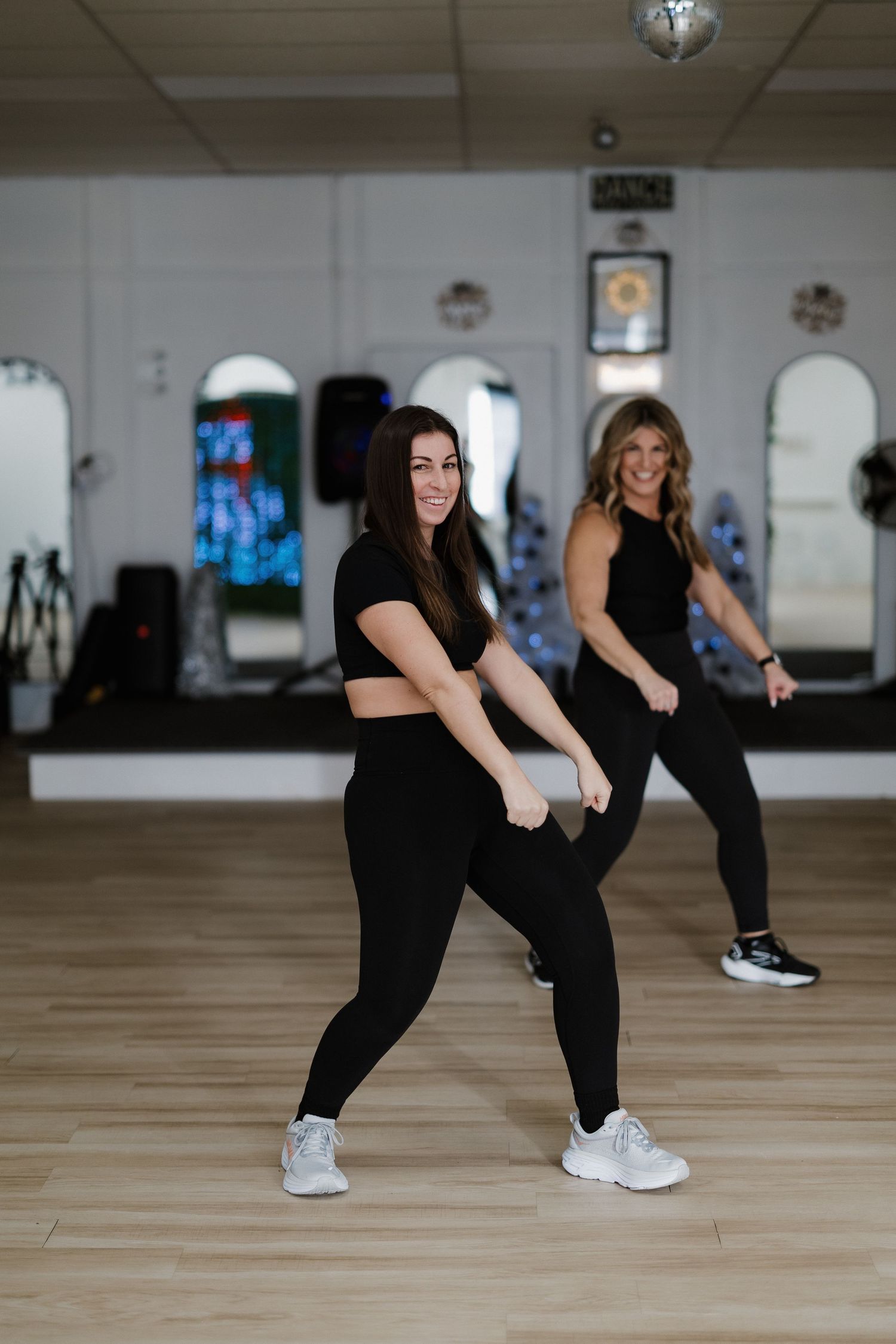 Two people in black workout attire doing dance fitness moves in a mirrored studio space.