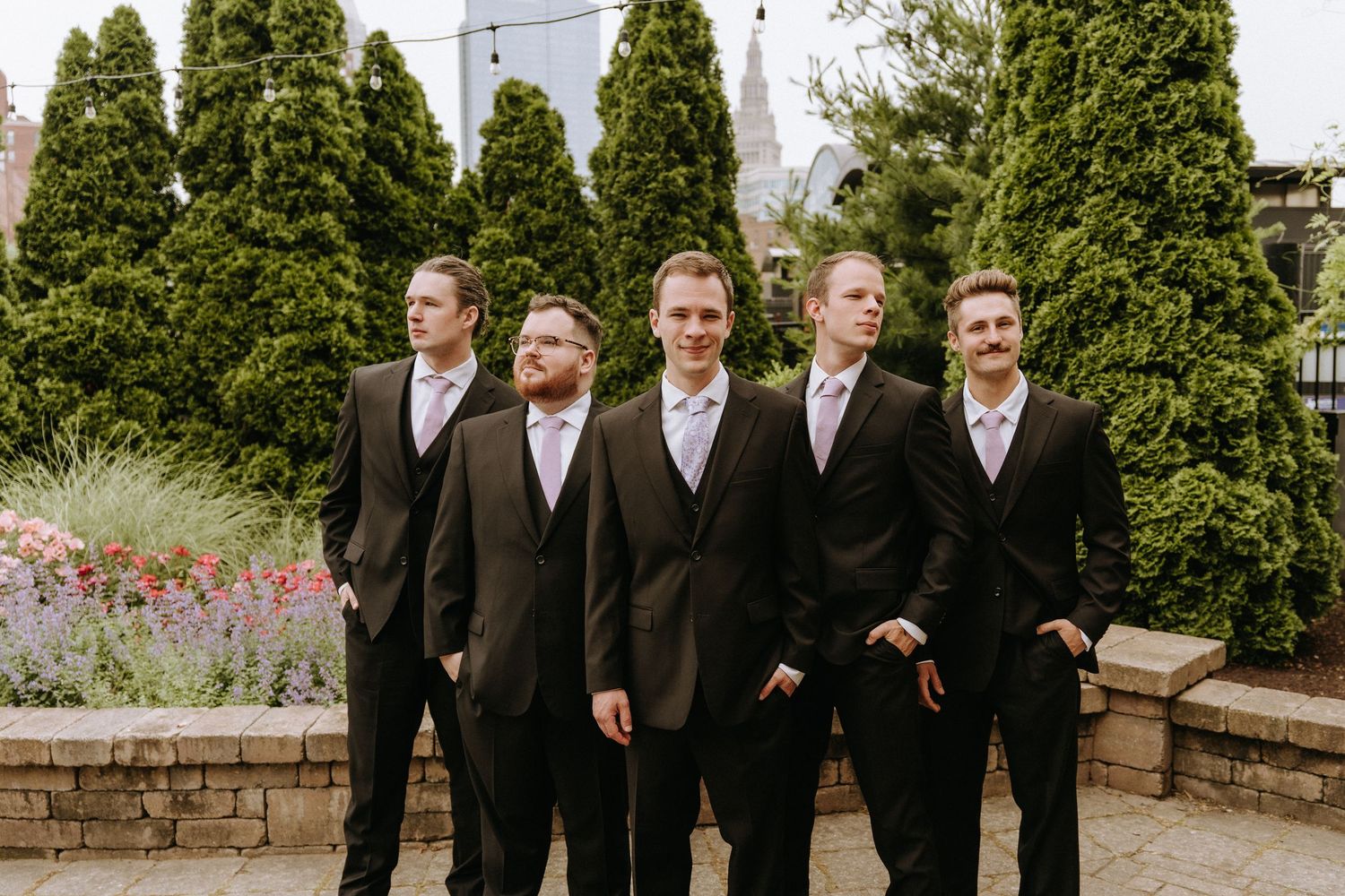 Groomsmen in black suits pose together outdoors with evergreen trees in the background.