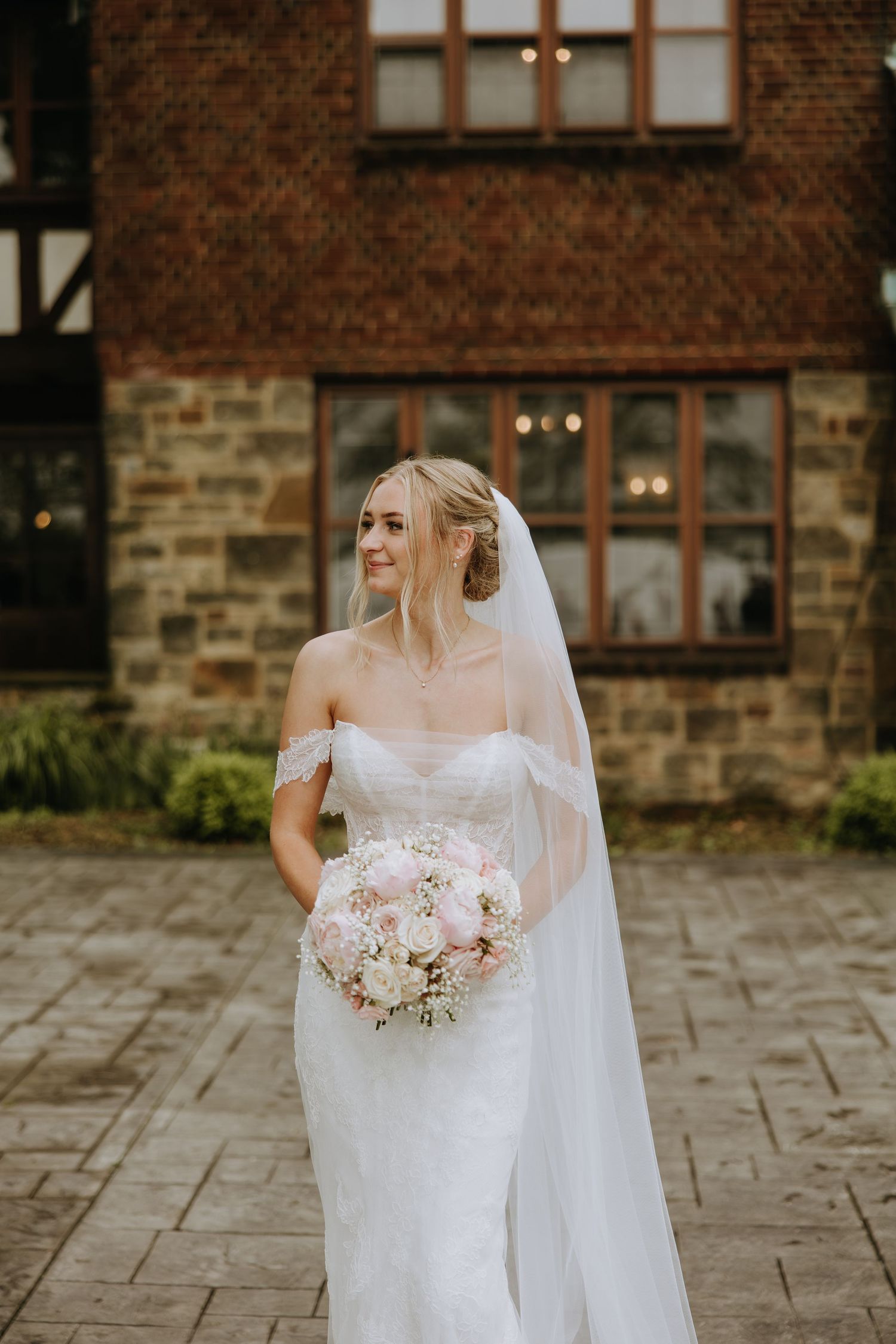 A bride in a white wedding dress holds a pink and white floral bouquet against a brick building backdrop.