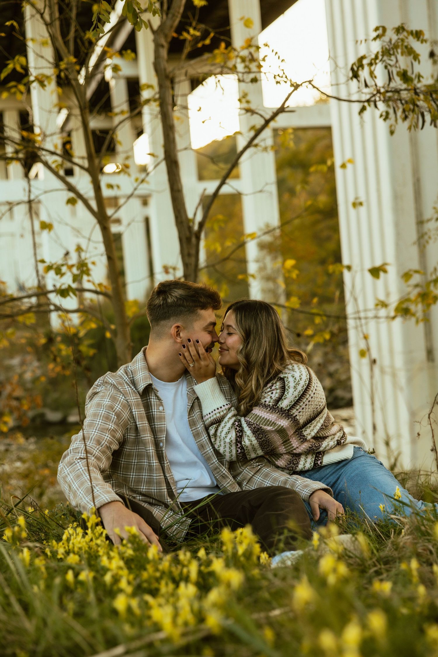 A couple shares a romantic moment while sitting in tall grass near white pillars during golden hour.