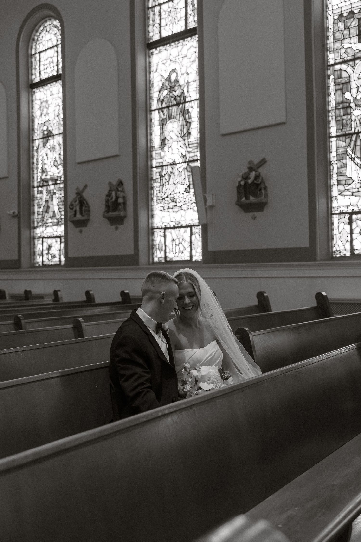 Black and white photo of couple sitting in church pew with stained glass windows.
