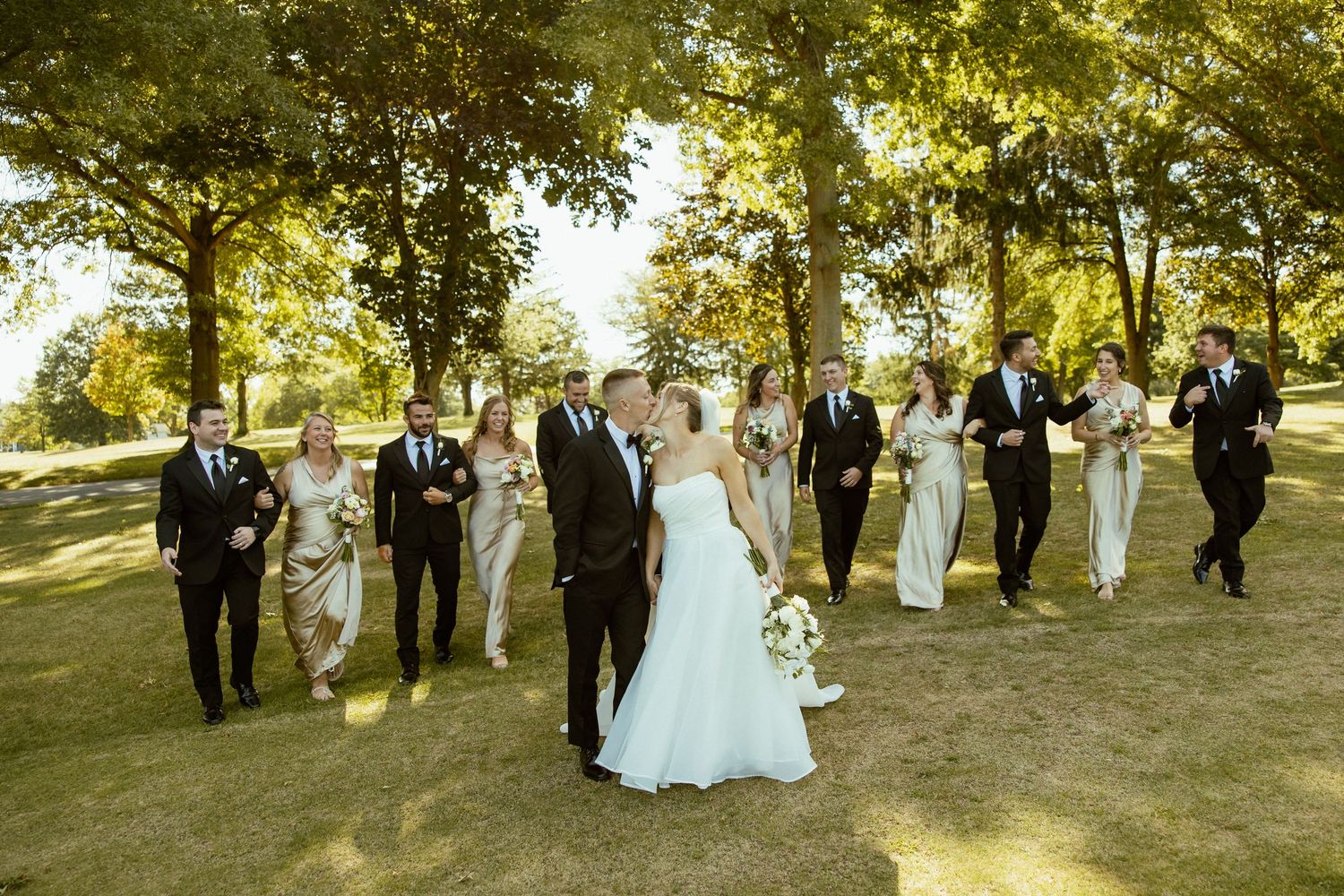 Wedding party walks together through a sunlit park with trees creating a warm autumn atmosphere.