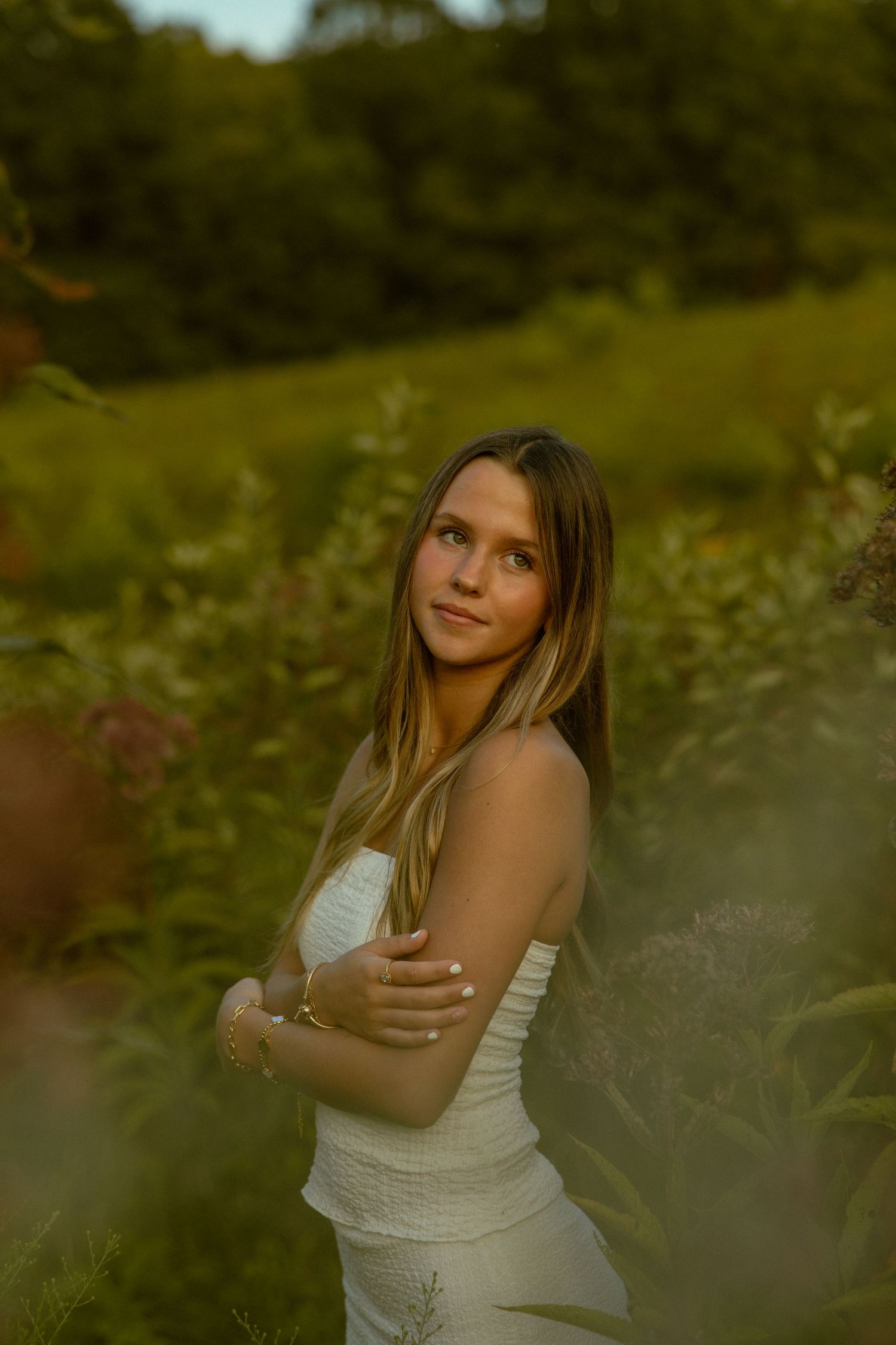 A person in a white tank top poses outdoors in a dreamy golden-hour setting among wildflowers and greenery.