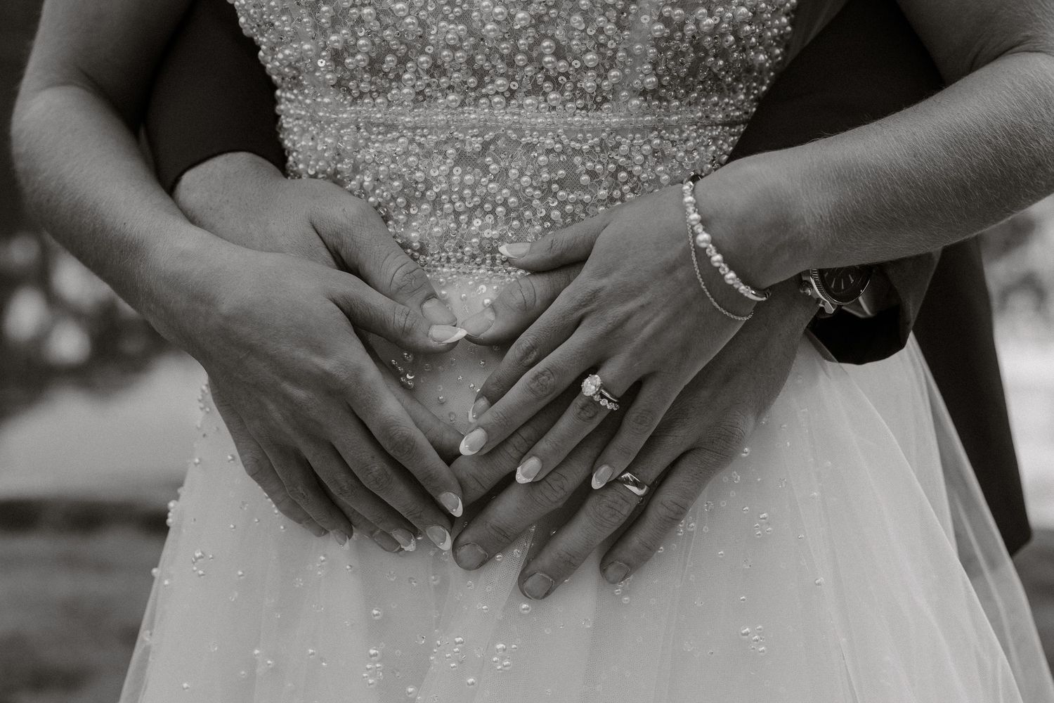 Black and white close-up of hands wearing wedding rings and bracelet resting on a tulle wedding dress.