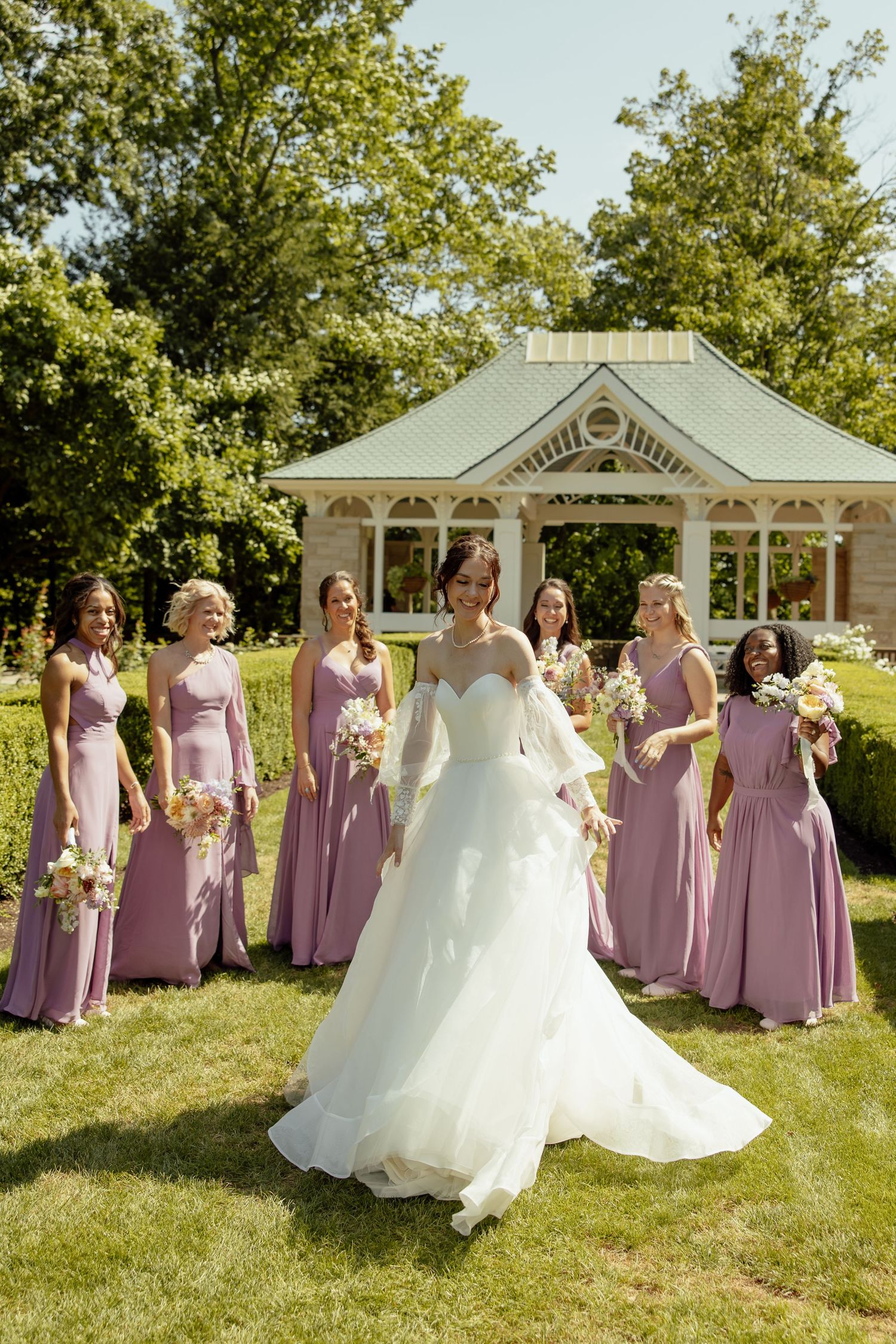 A wedding party in lavender bridesmaid dresses poses on a sunny lawn with a white gazebo in the background.