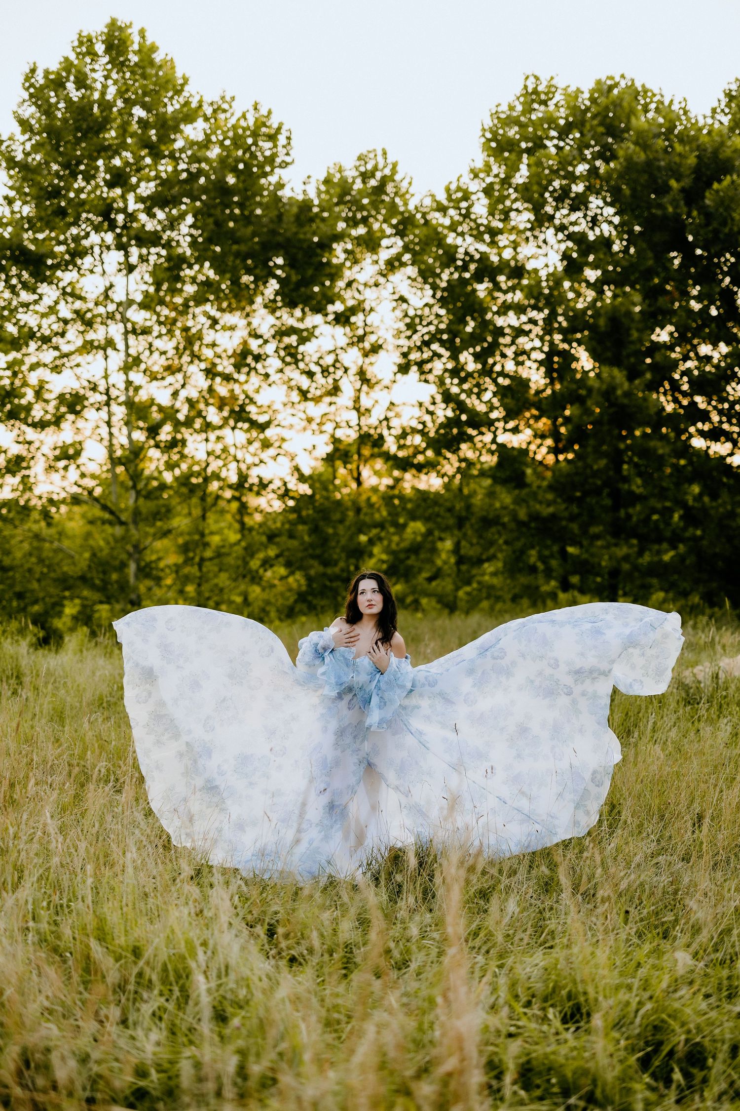 An ethereal white dress billows in the breeze against a backdrop of trees at sunset.