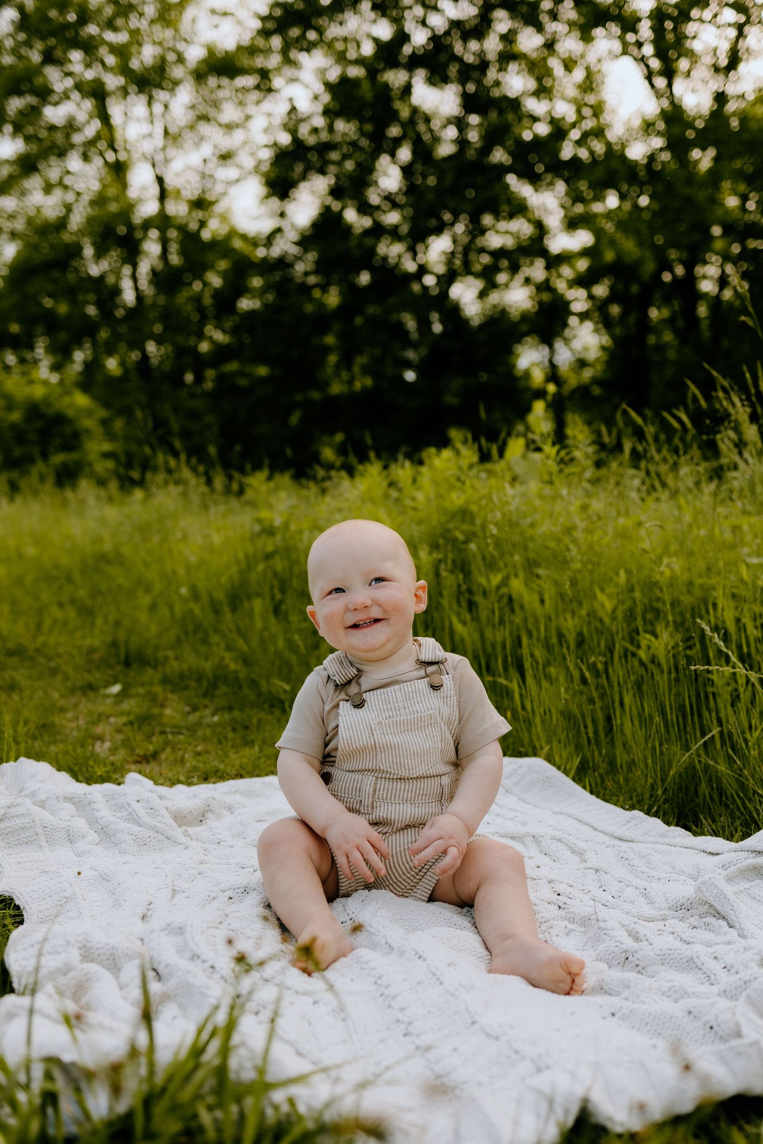 A smiling baby in tan overalls sits on a white blanket in a lush green outdoor setting.