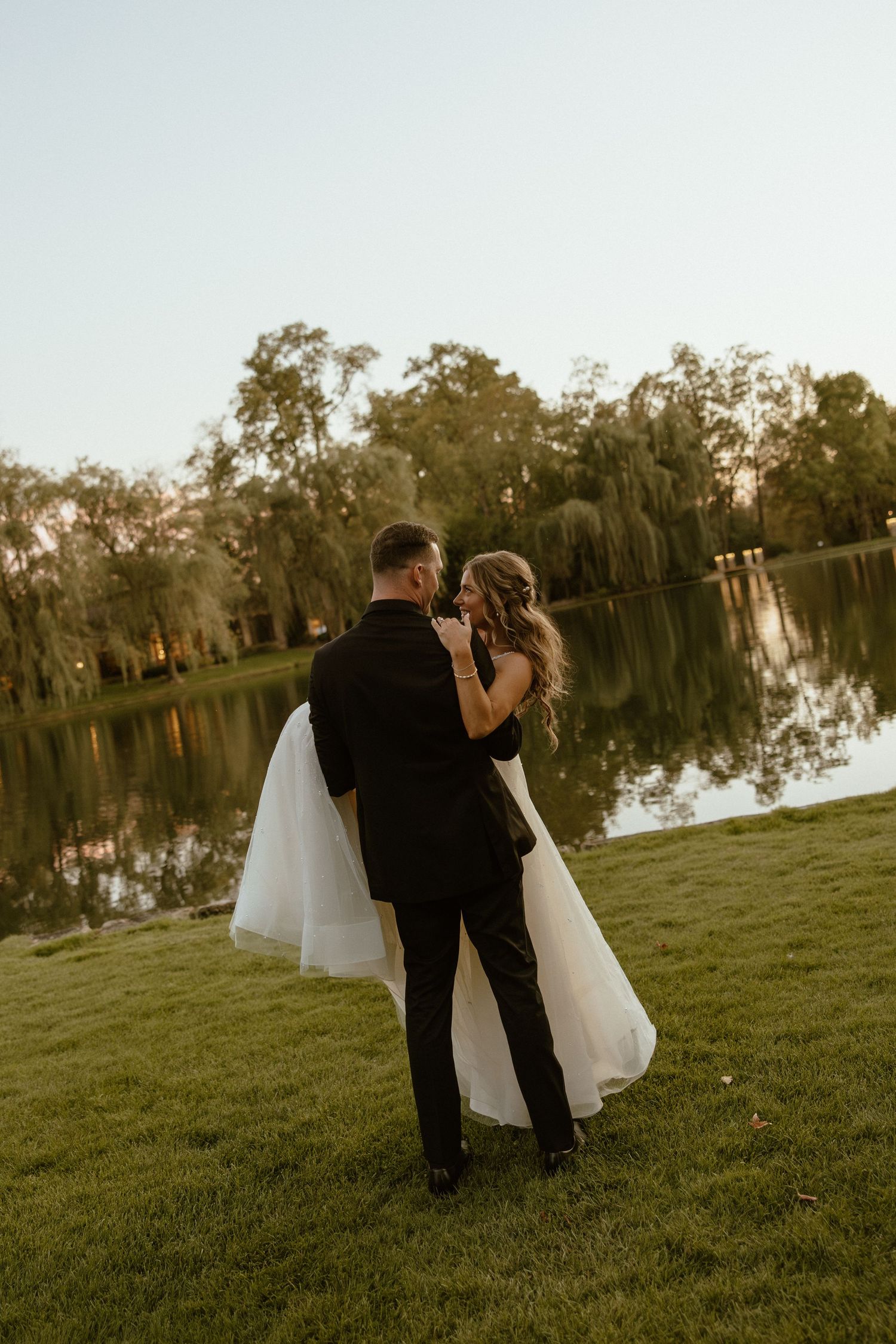 A romantic wedding couple embraces on a grassy lawn beside a tranquil lake at sunset.